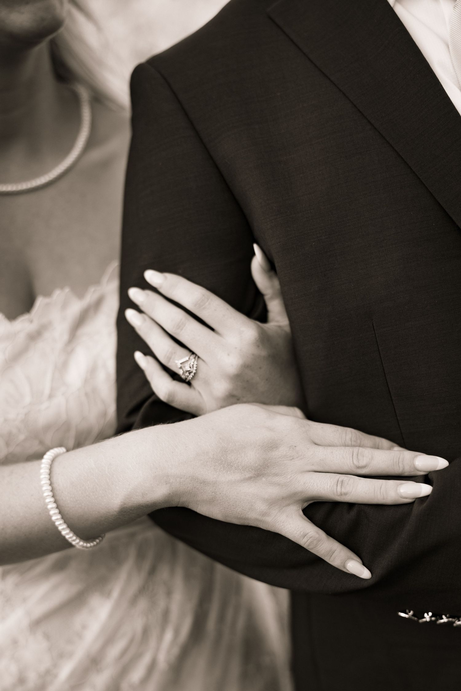Black and white close-up of bride's hands with diamond ring and pearl bracelet on groom's suit.
