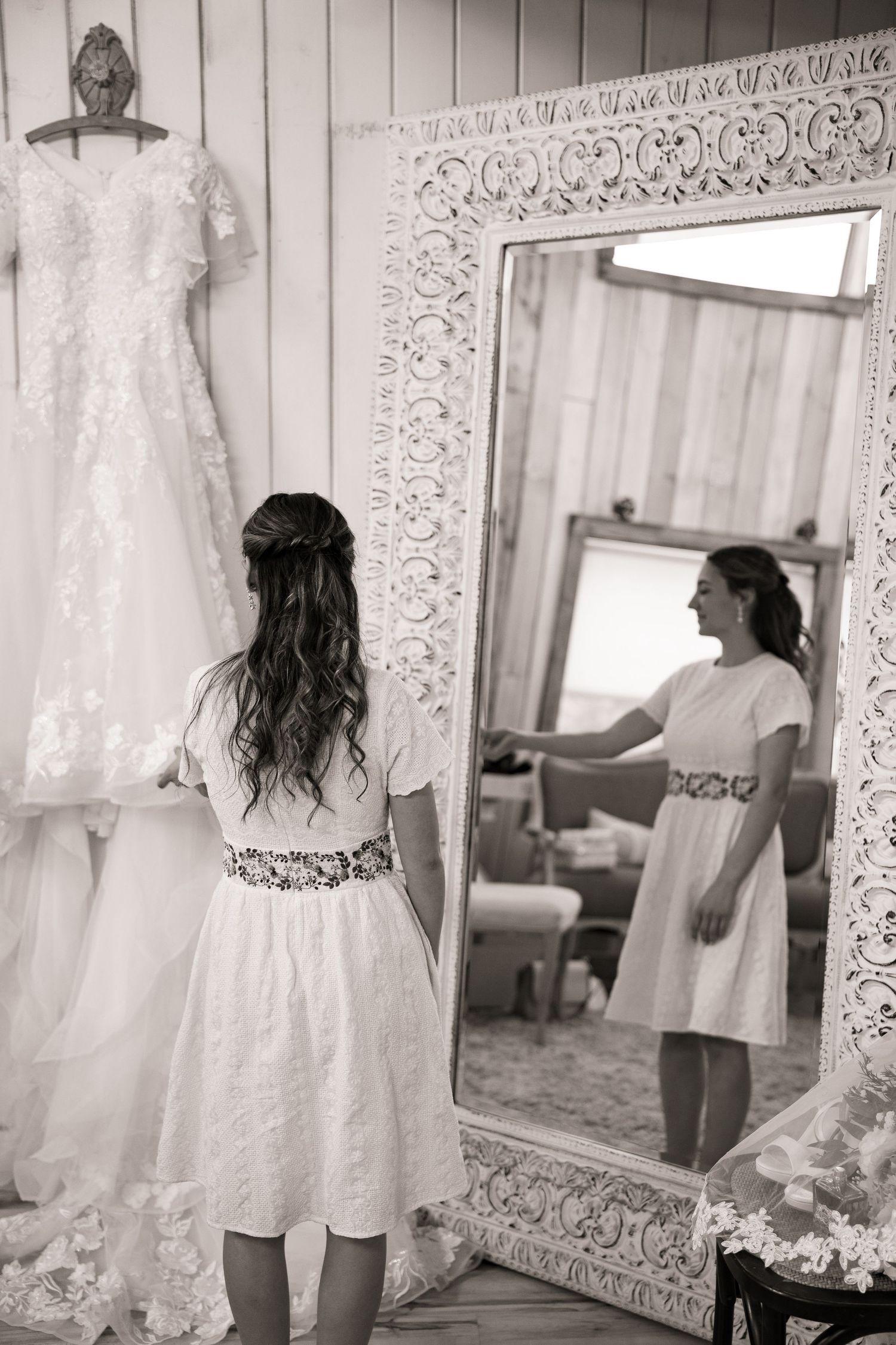 Black and white photo of bride in white dress looking at her reflection in ornate mirror.
