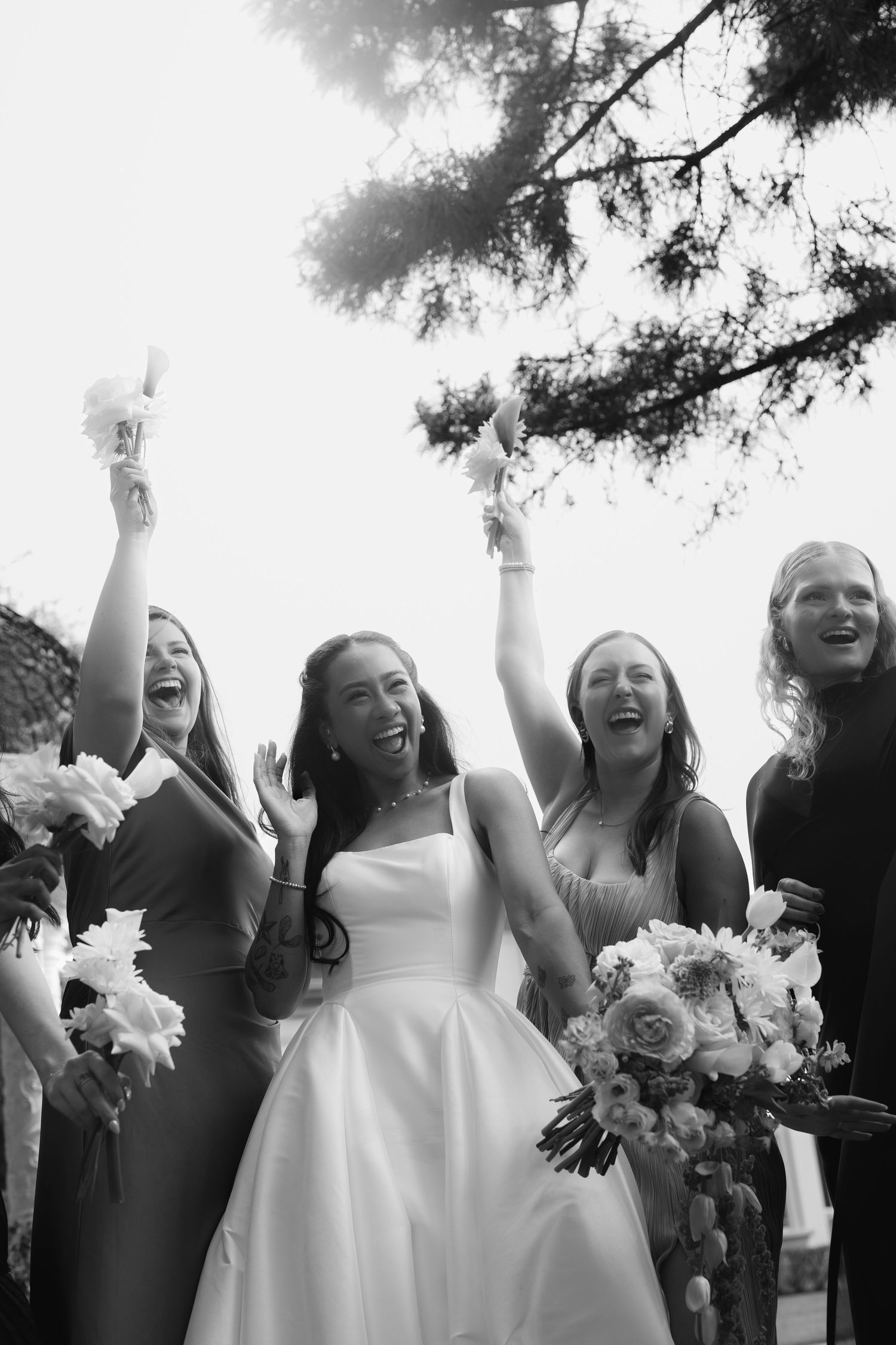 Black and white photo of joyful bride and bridesmaids celebrating outdoors with raised bouquets and laughter.