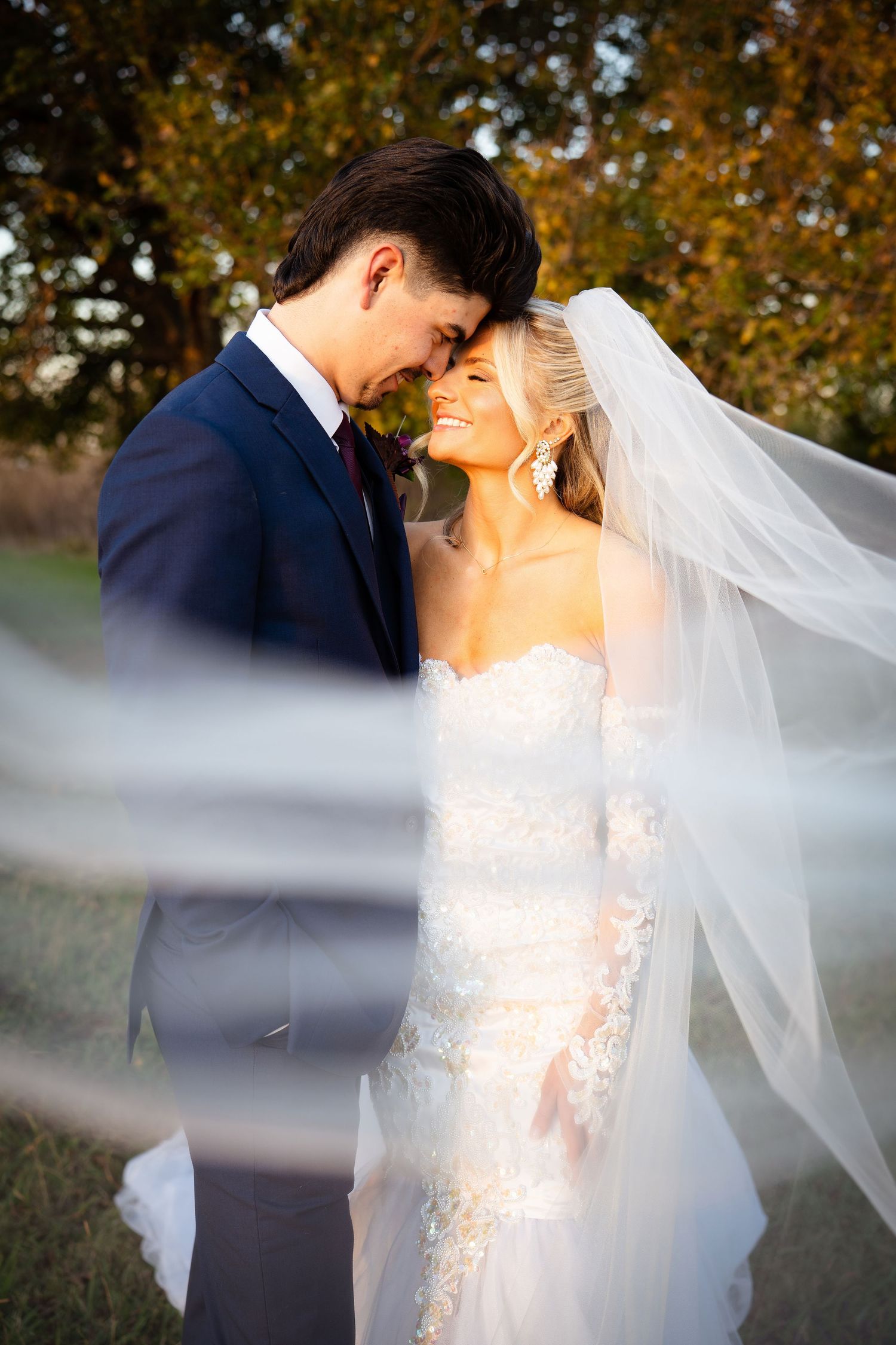 Bride and groom share a romantic forehead-to-forehead moment as her veil billows in the breeze outdoors.