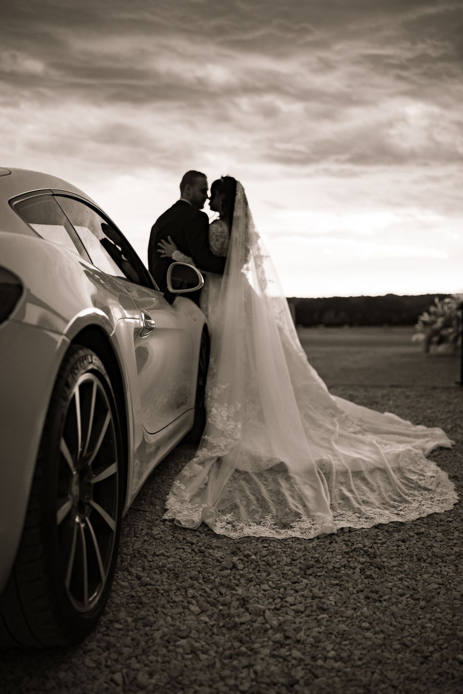 Romantic black and white photo of bride and groom kissing beside a luxury car.