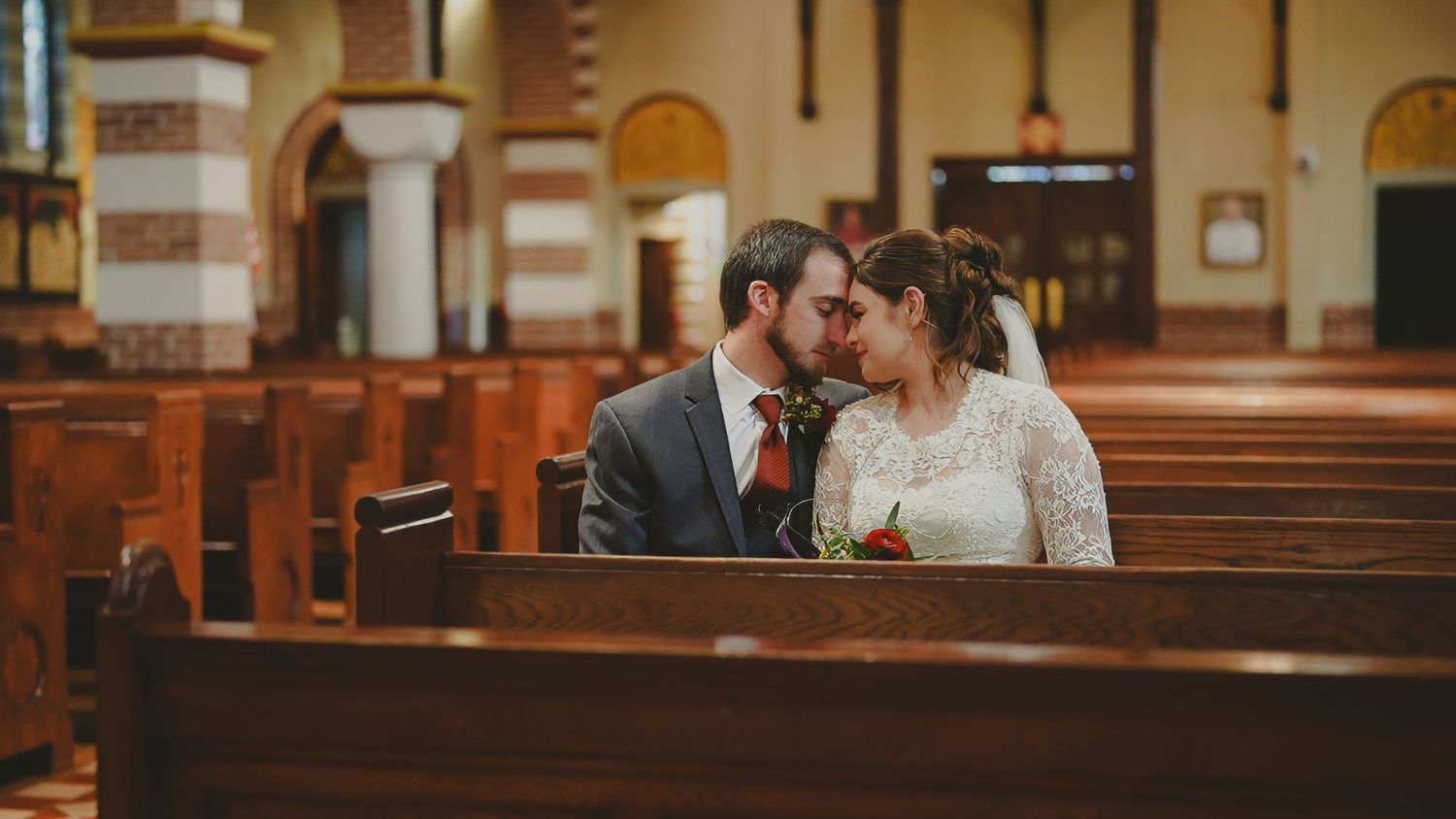 Bride and groom share a tender kiss while seated in a wooden church pew during their wedding ceremony.