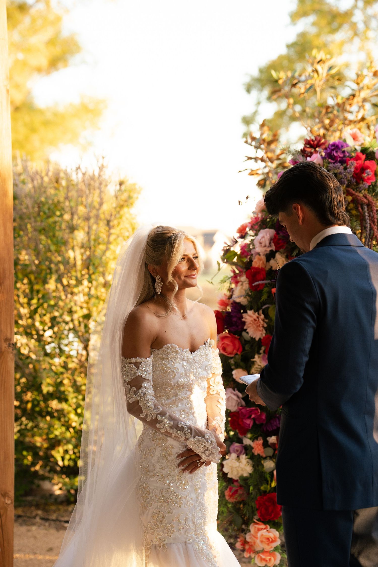 Radiant bride smiling at groom during outdoor wedding ceremony beside vibrant colorful floral arch.