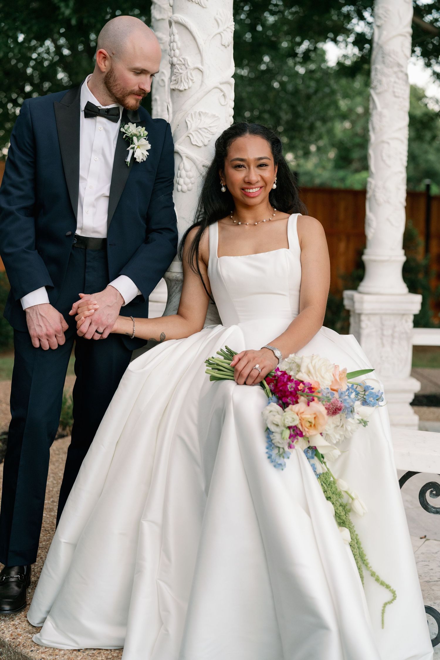 A bride in a white ballgown holds a colorful bouquet beside her groom in a navy tuxedo outdoors.