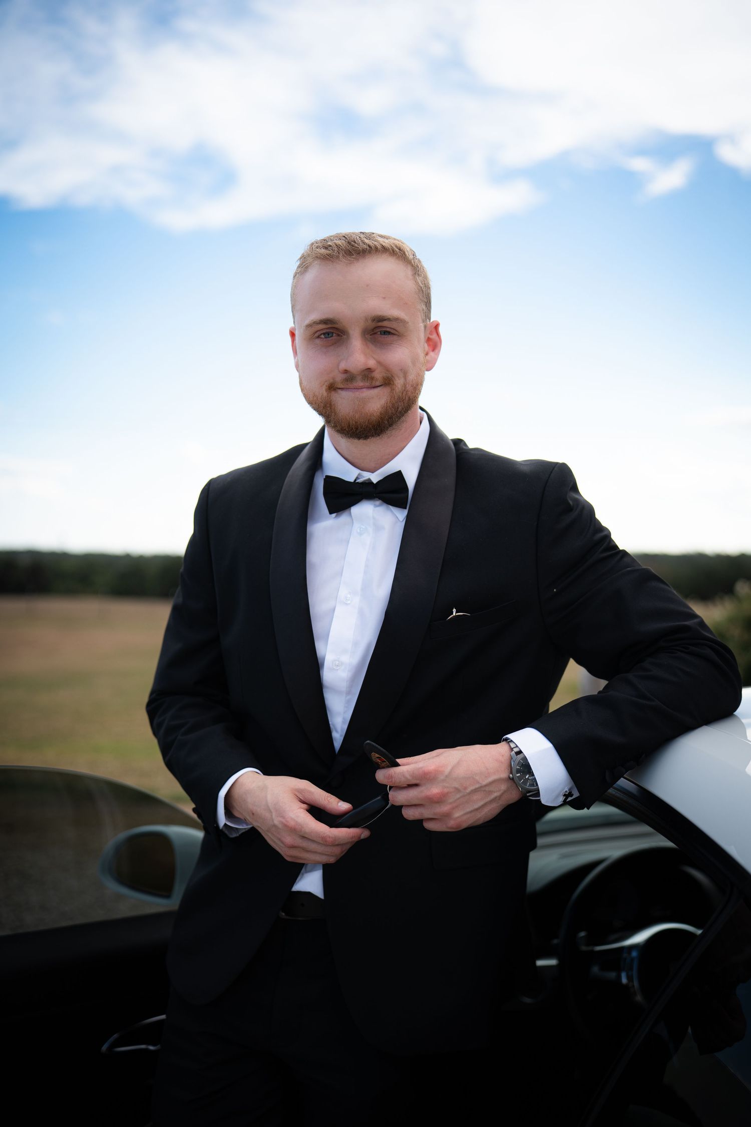 Dapper groom in black tuxedo with bow tie leaning against white car outdoors.