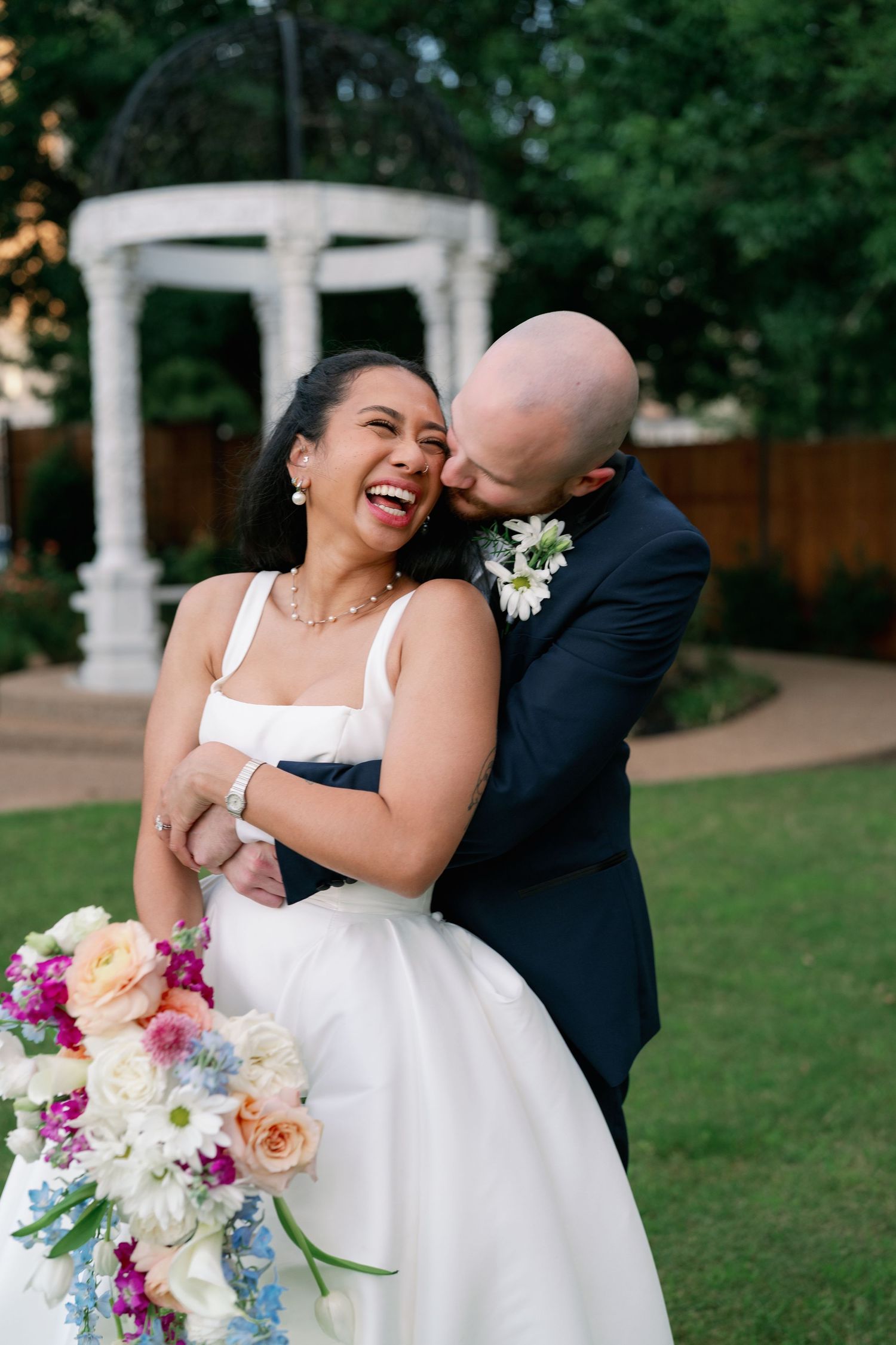 A laughing bride and bald groom share a joyful embrace outdoors near a white garden gazebo.