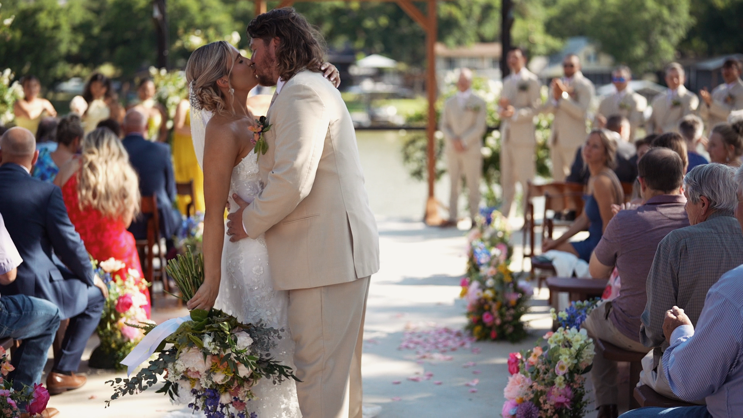 A bride and groom share their first kiss at an outdoor wedding ceremony lined with colorful floral arrangements.
