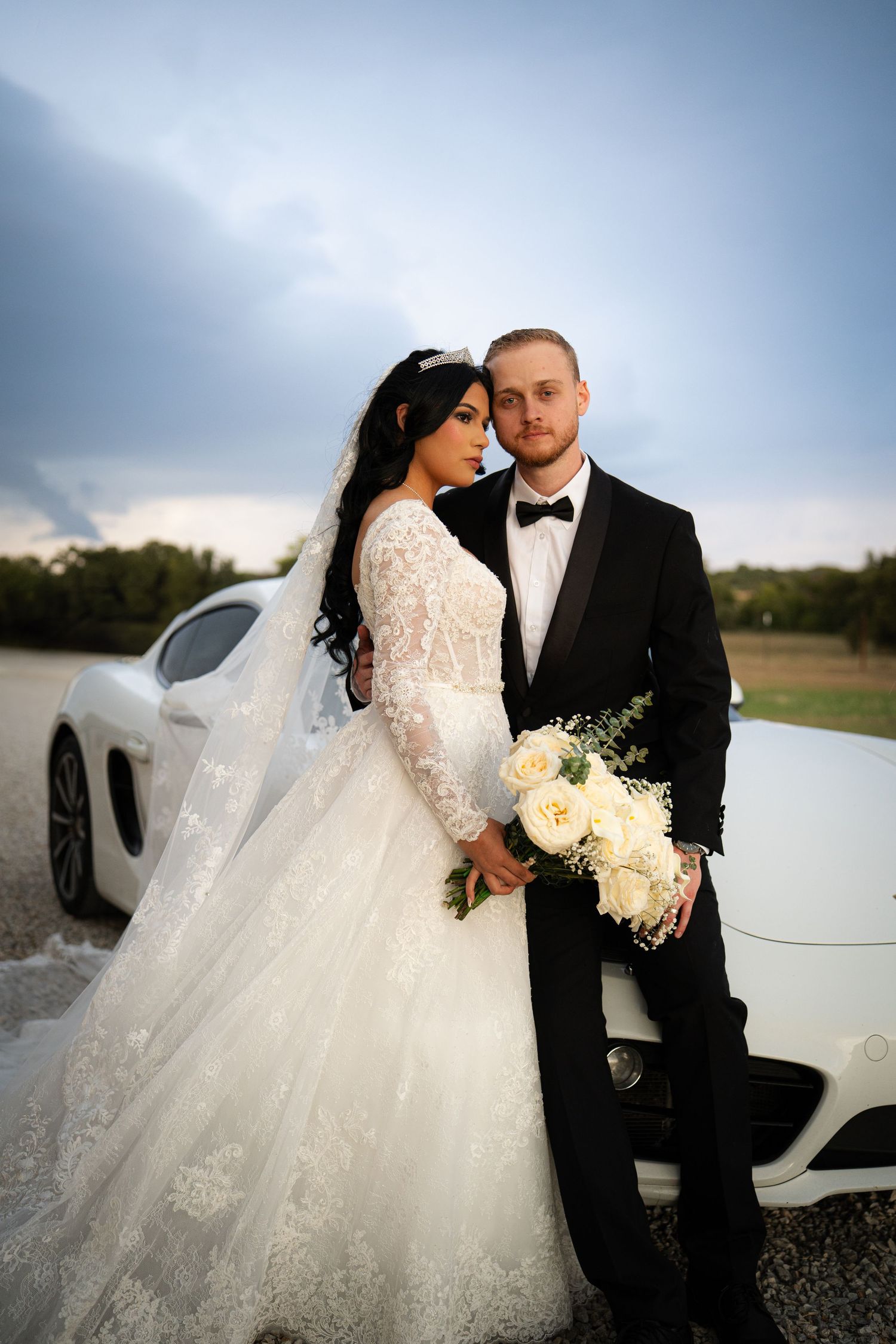 Bride and groom posing elegantly beside a white Porsche sports car at sunset.