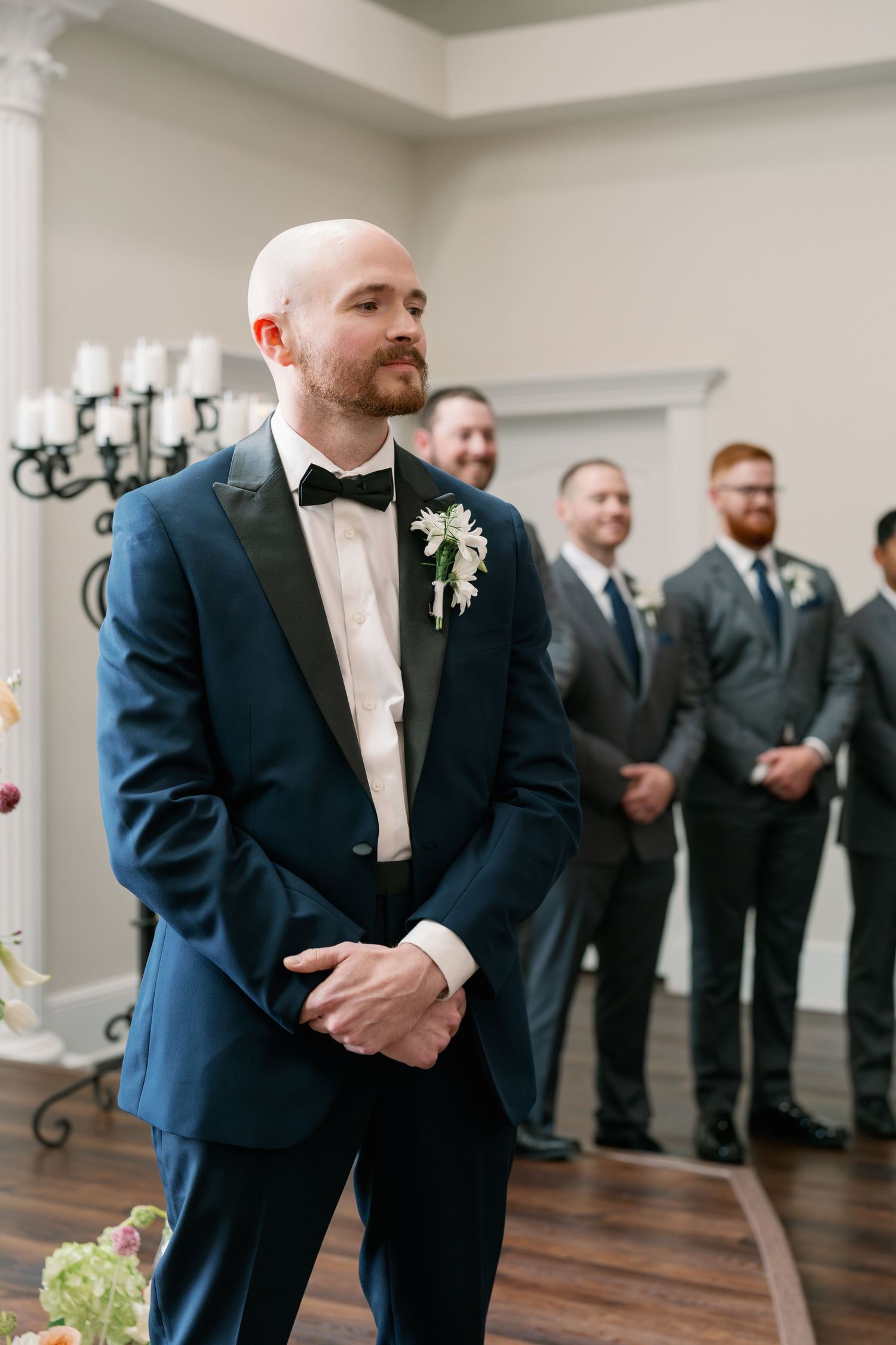 A groom in a navy tuxedo waits at the altar with groomsmen standing behind him at a wedding.