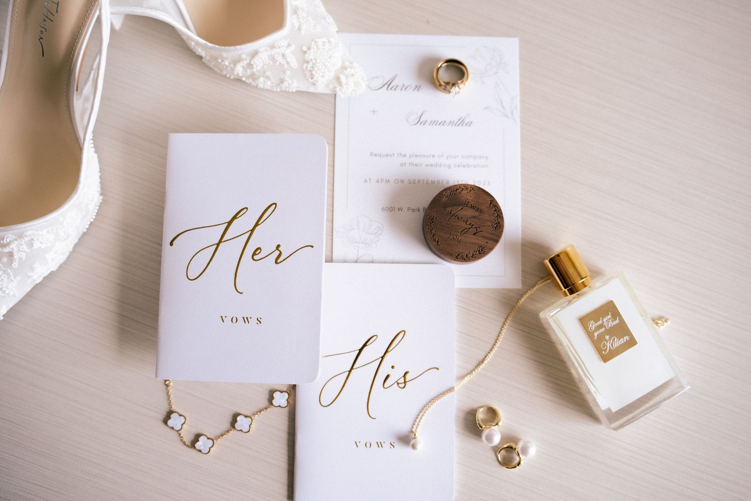 A wedding flat lay displaying gold-lettered vow books, invitation, perfume, bridal heels, and delicate jewelry accessories.