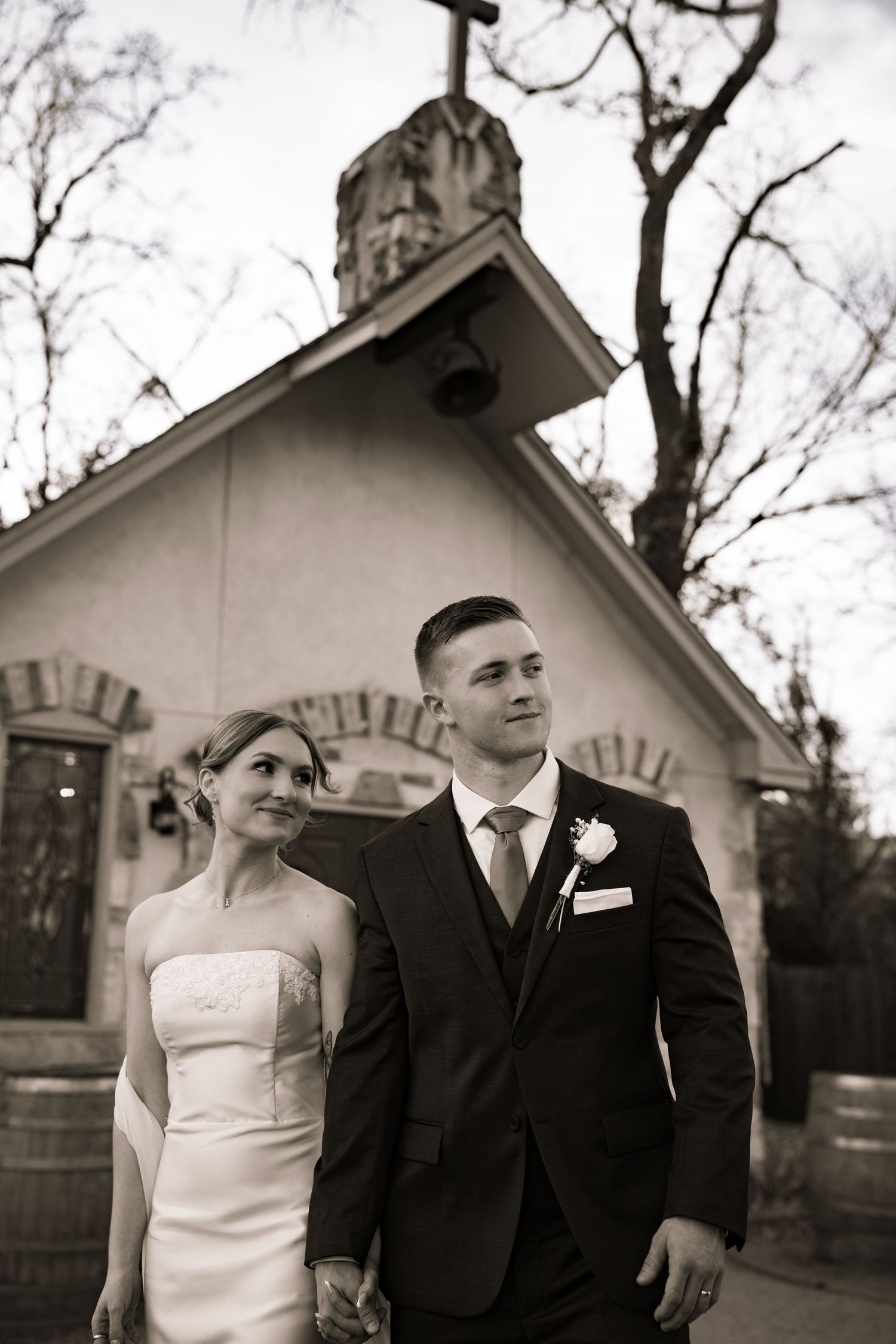 Black and white portrait of newlyweds holding hands outside rustic chapel at dusk.