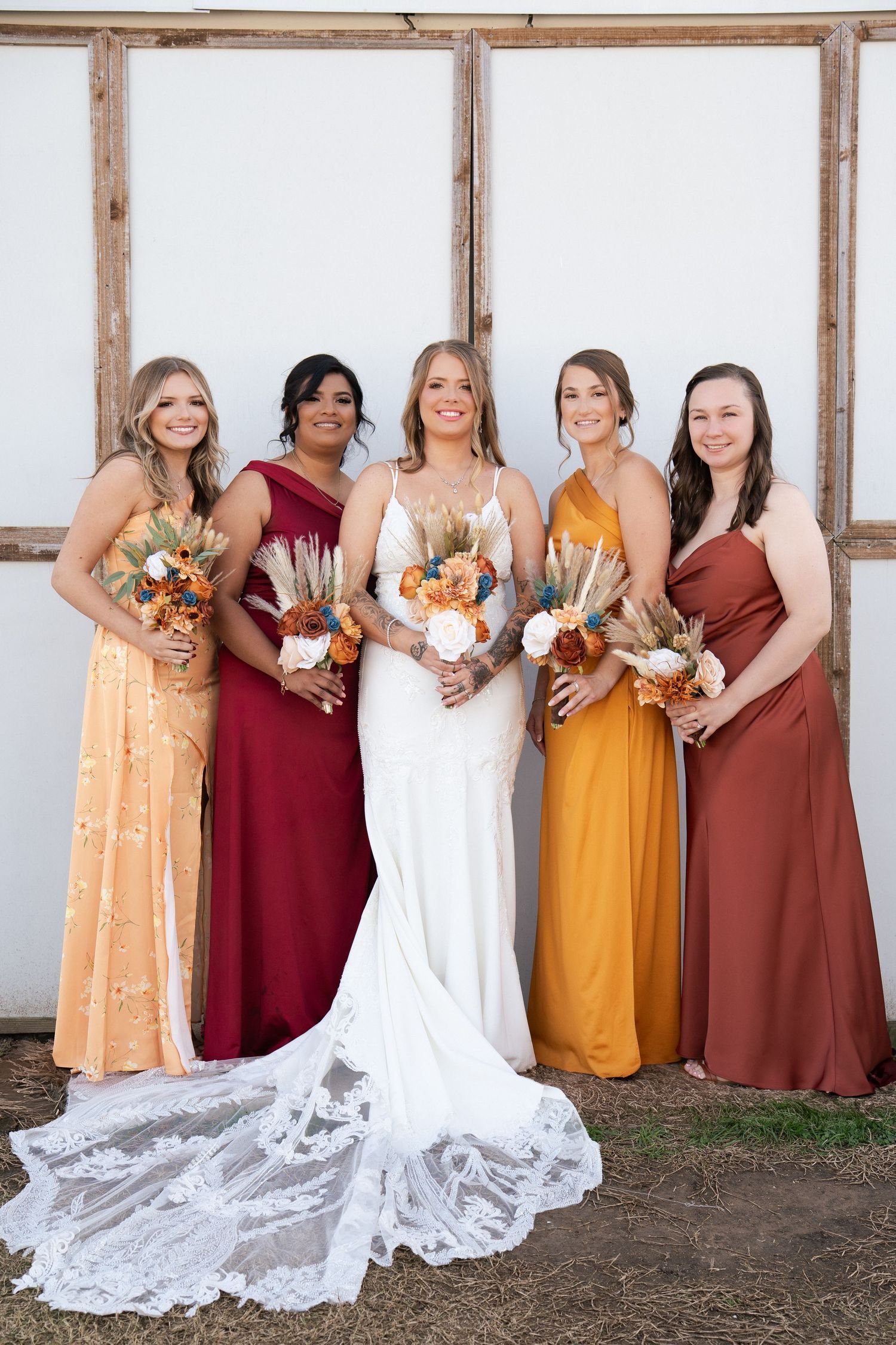 Bride in white gown with four bridesmaids in autumn-toned dresses holding rustic bouquets outdoors.