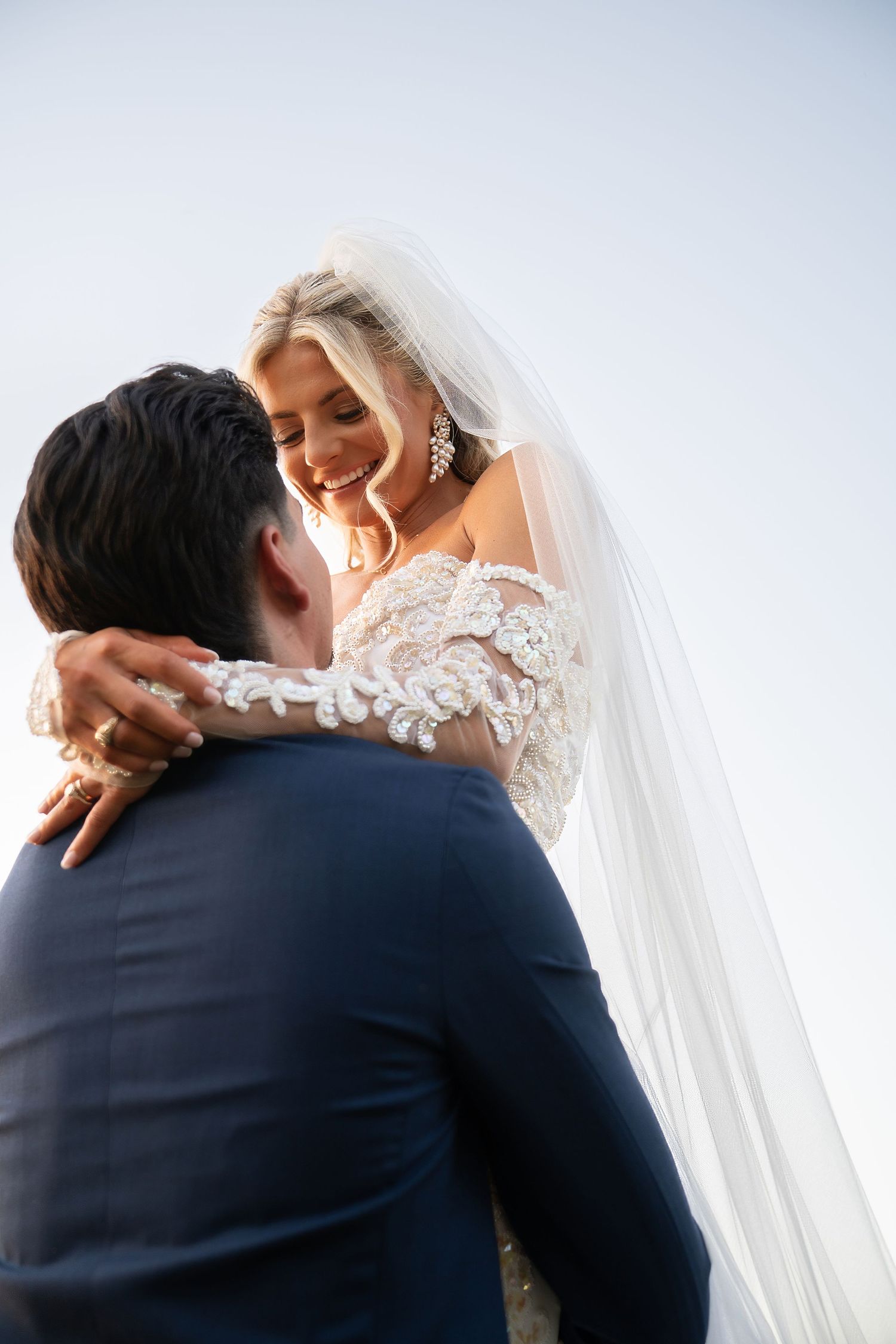 Radiant bride in lace gown laughs joyfully while embracing groom against a clear blue sky backdrop.