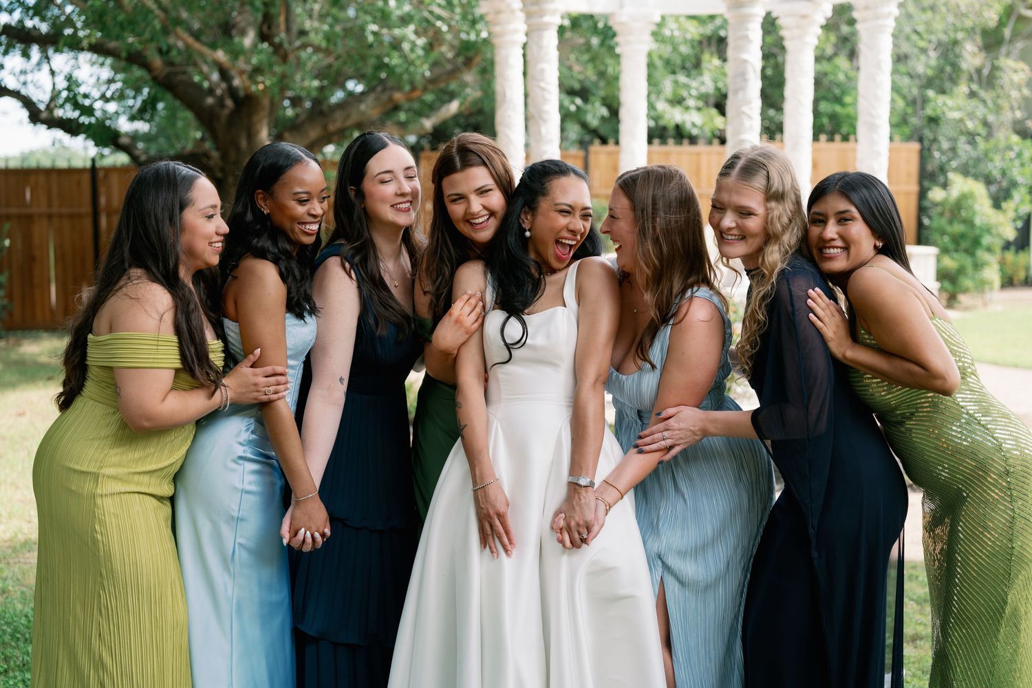 Bride laughing with bridesmaids in colorful mismatched dresses outdoors at wedding celebration.