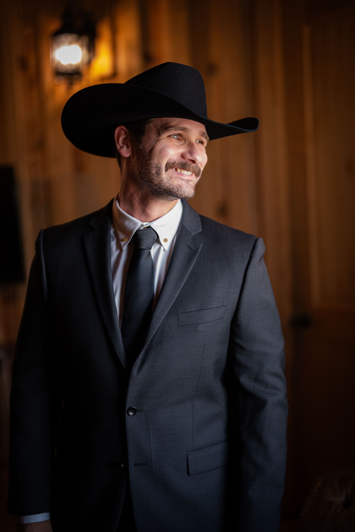 Smiling man in dark suit, tie, and black cowboy hat standing in warmly lit rustic wooden interior.