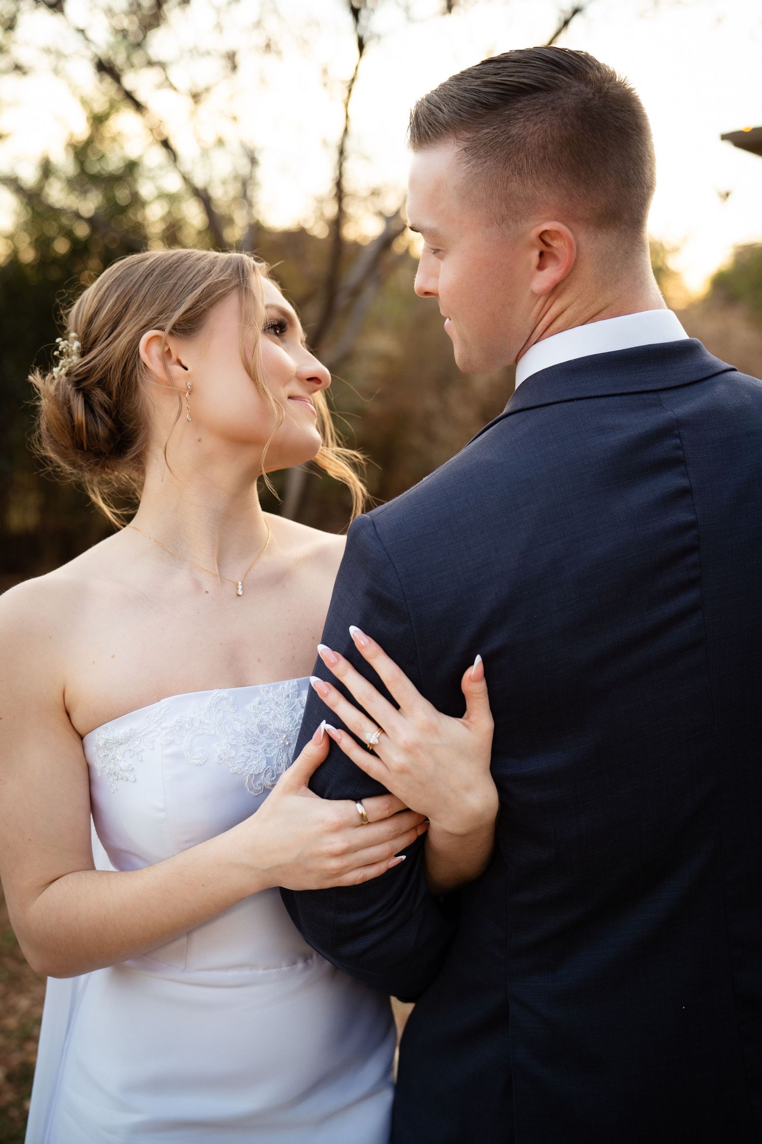 Bride in strapless white gown smiles up at groom in navy suit during golden hour wedding portrait.