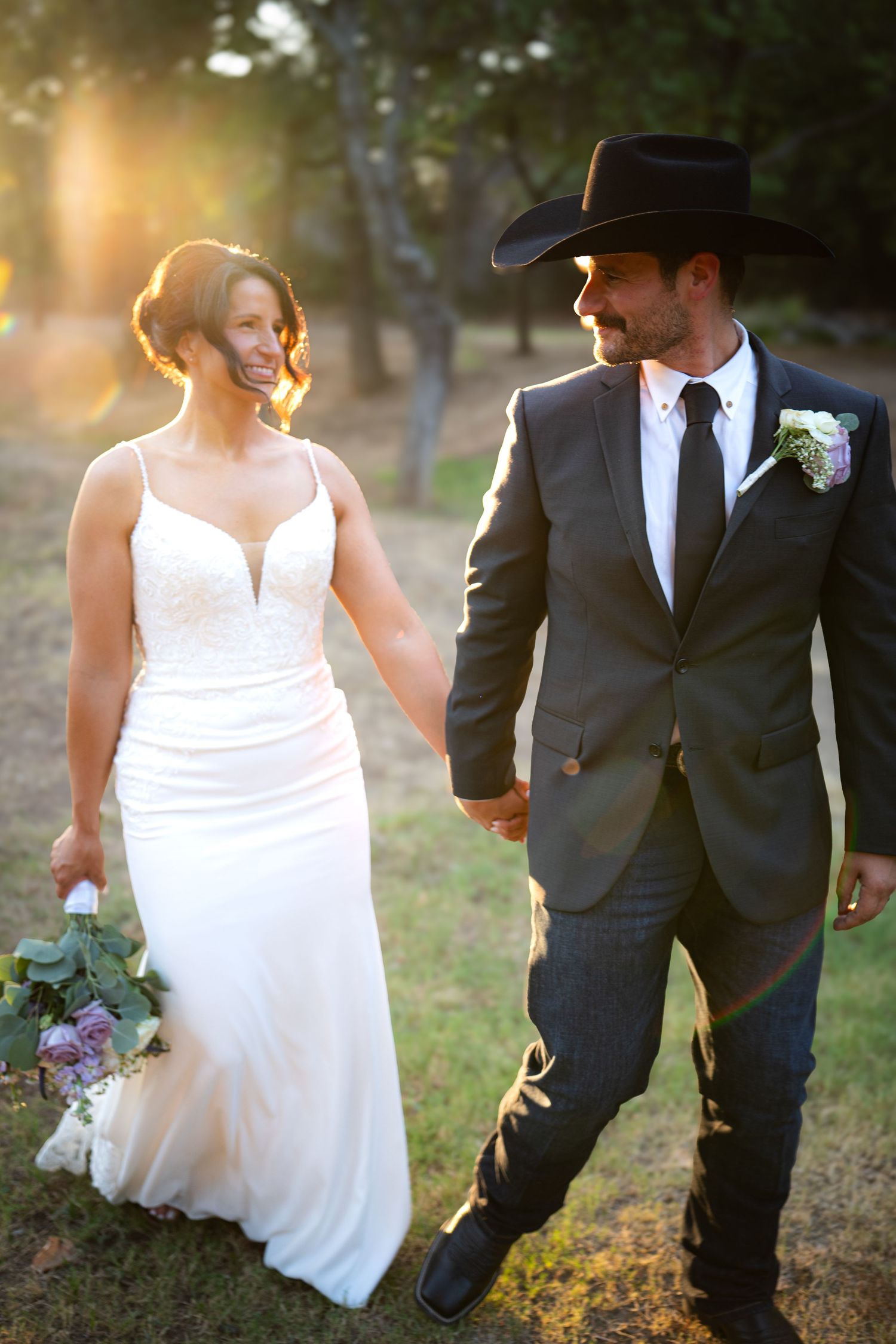 Bride and groom holding hands at golden hour, she in white gown with bouquet, he in gray suit and black cowboy hat.
