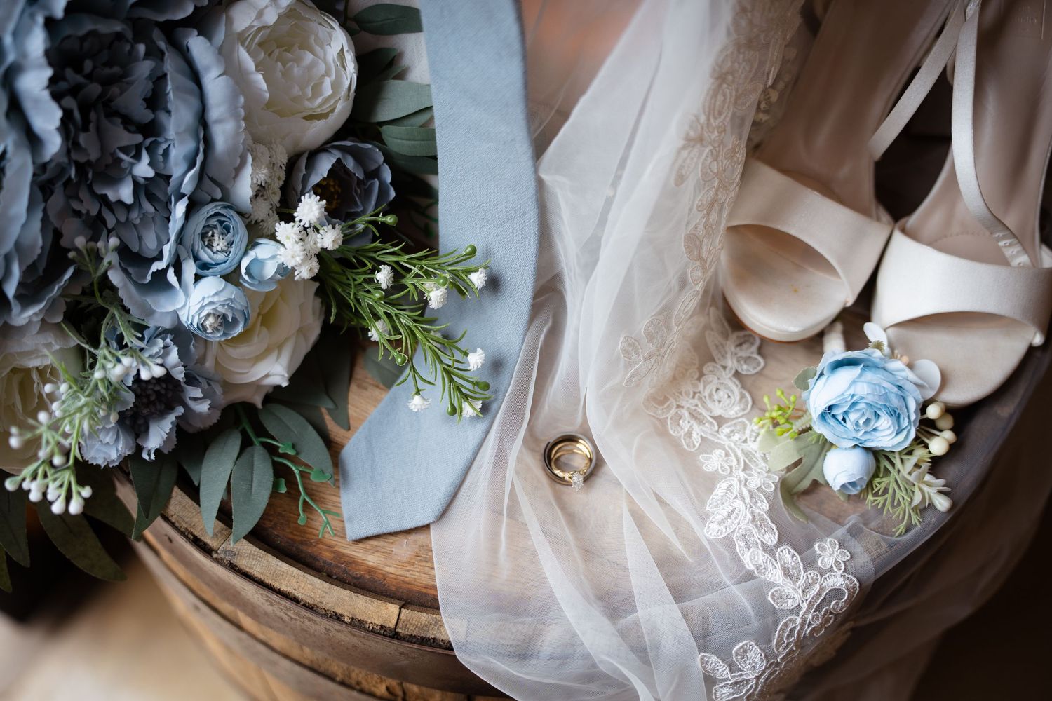 Wedding flat lay with blue floral bouquet, bridal heels, veil, boutonniere, and gold wedding band.