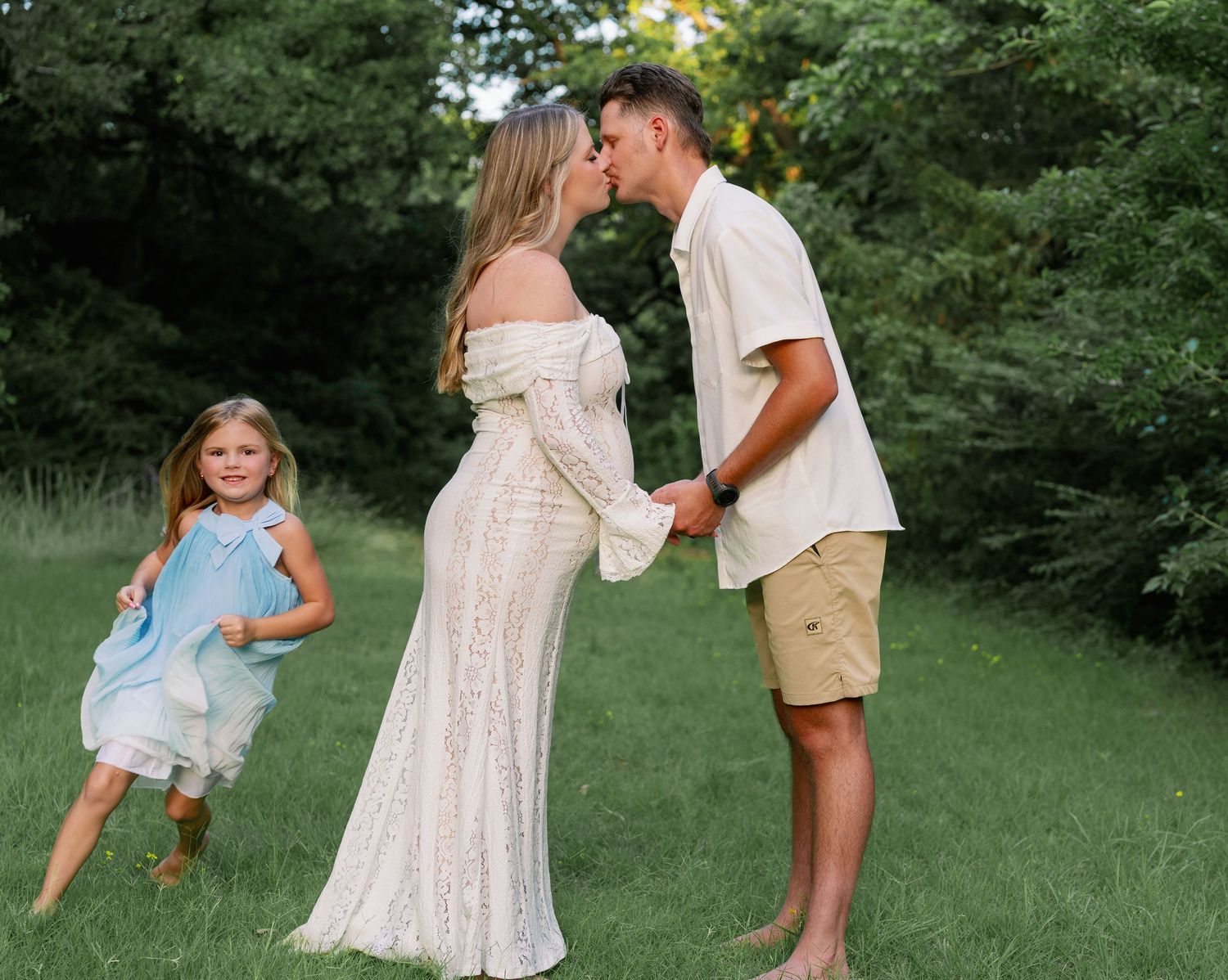 Expecting mother in white lace gown kisses partner while young daughter twirls nearby in green outdoor setting.