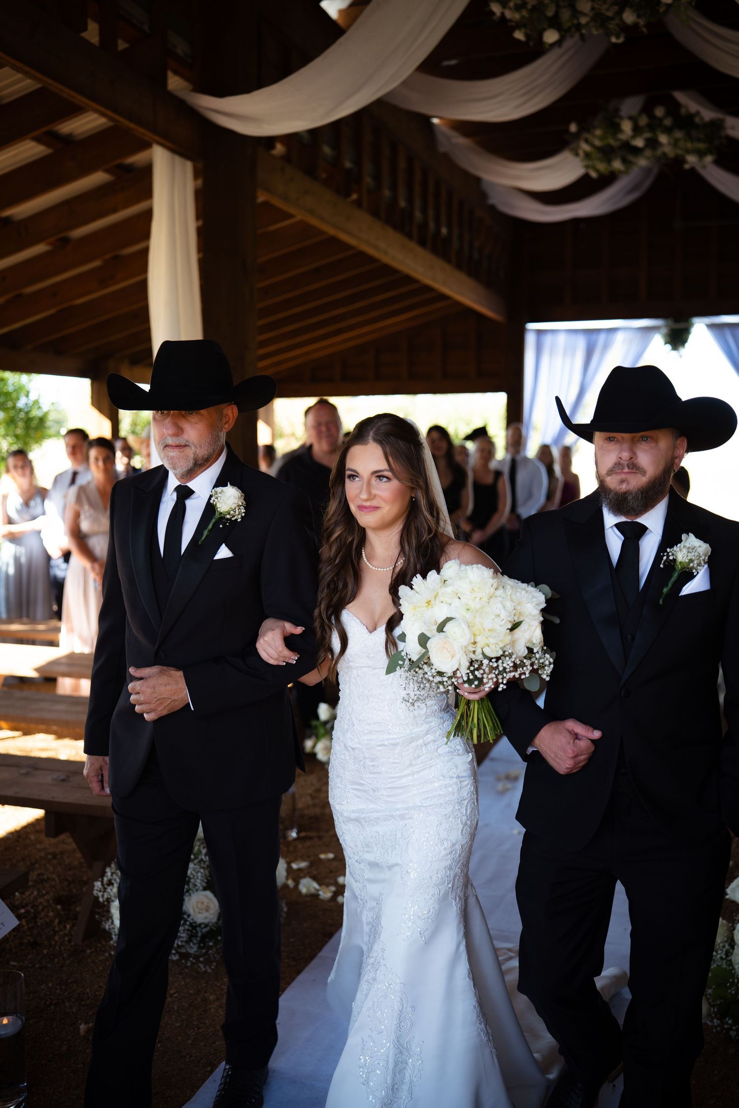 Bride escorted down the aisle by two men in black suits and cowboy hats at rustic wedding.