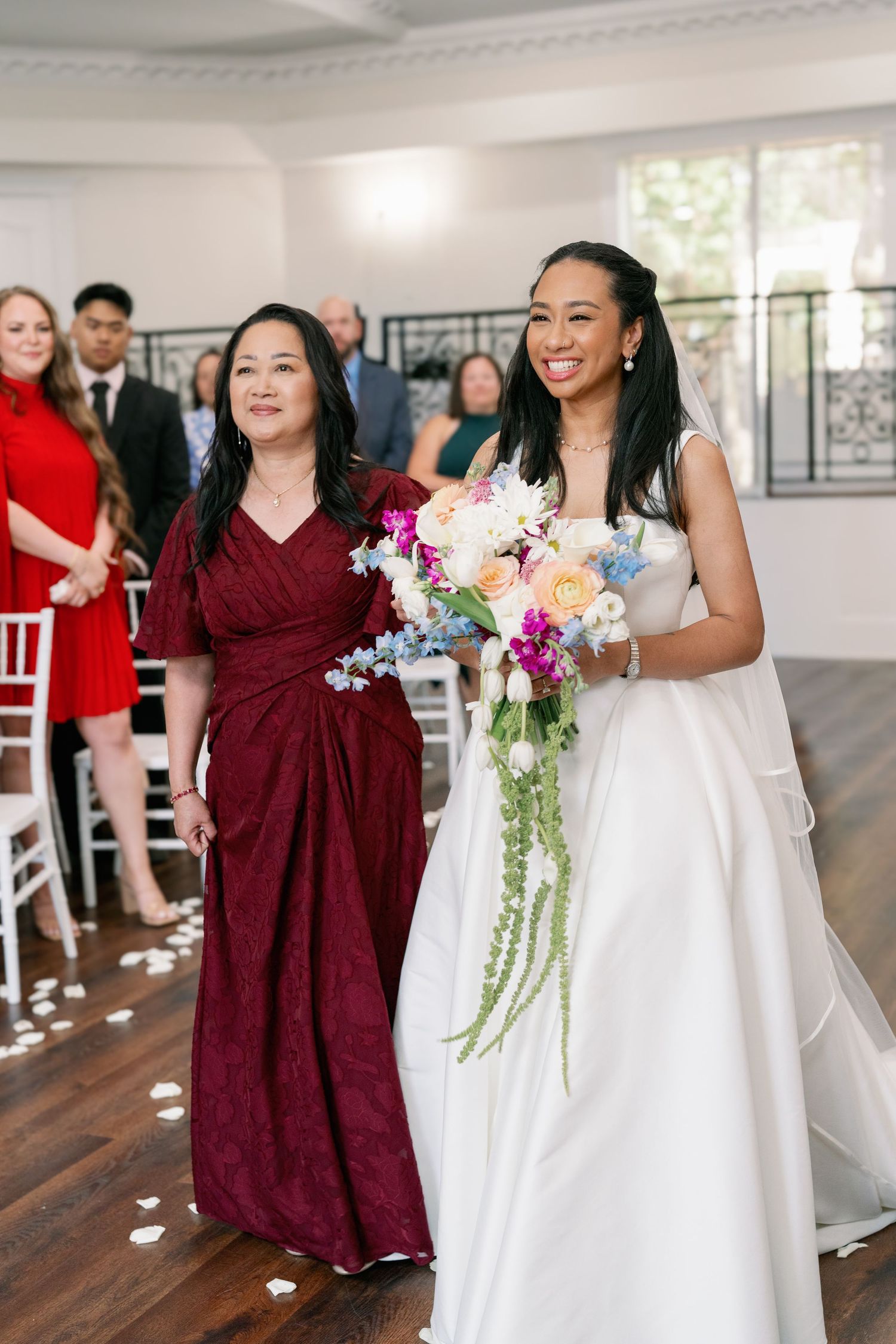 A beaming bride in a white gown walks down the aisle with her mother during a wedding ceremony.