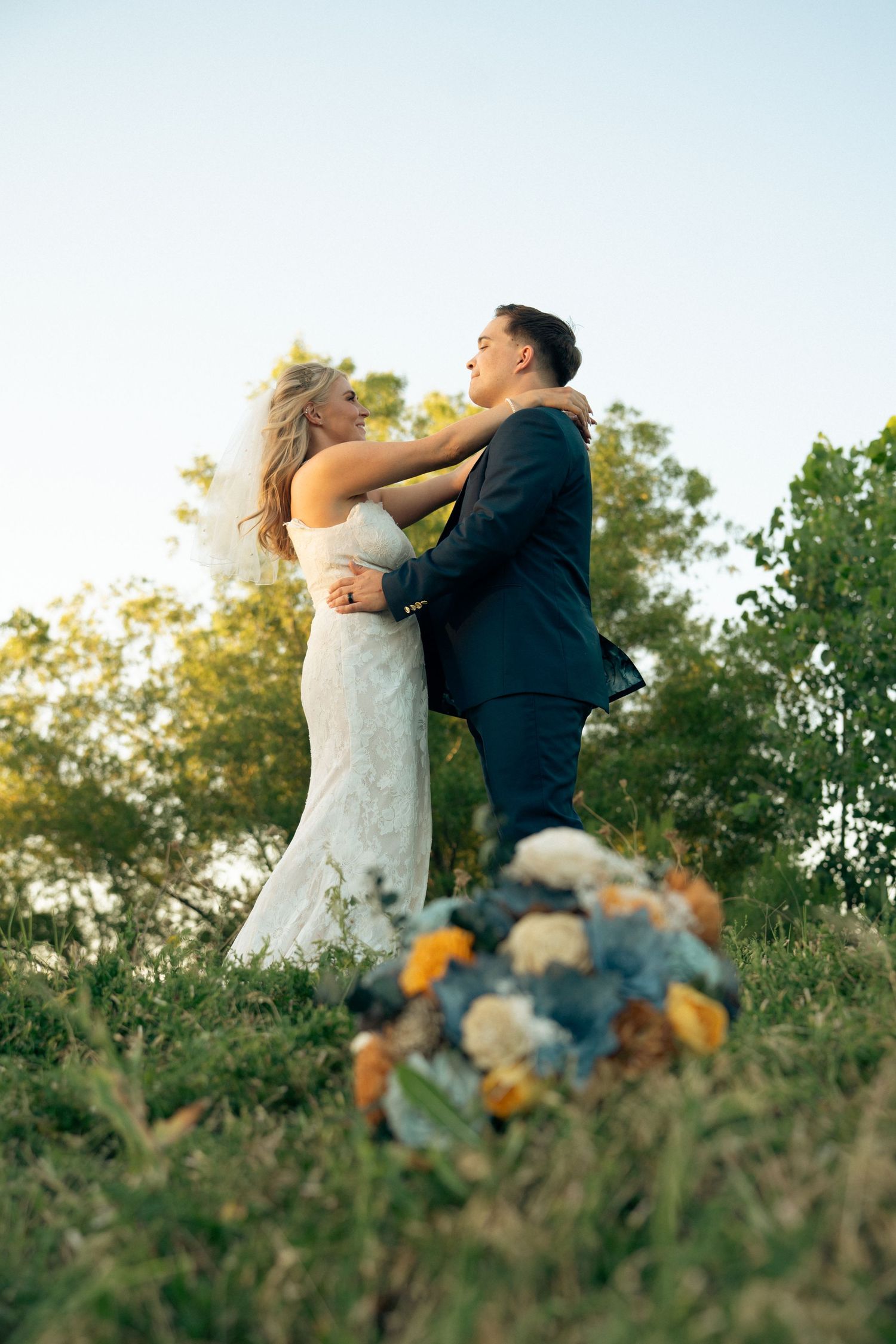 Bride and groom embracing on grassy hill at sunset with colorful bouquet in foreground.