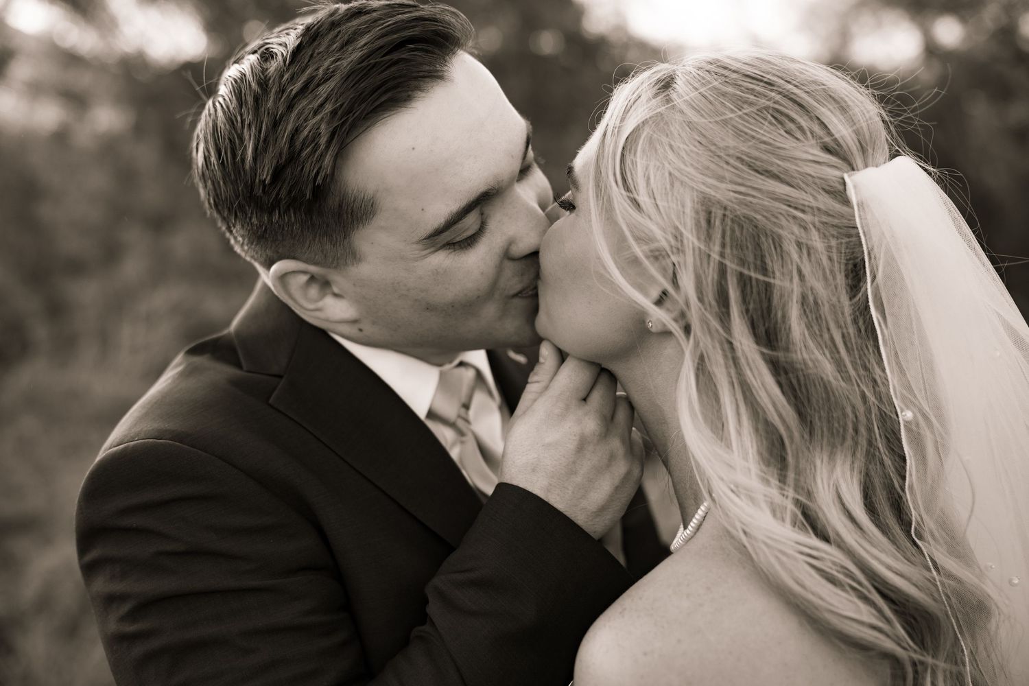 A bride and groom share a romantic kiss in a classic black and white wedding portrait outdoors.