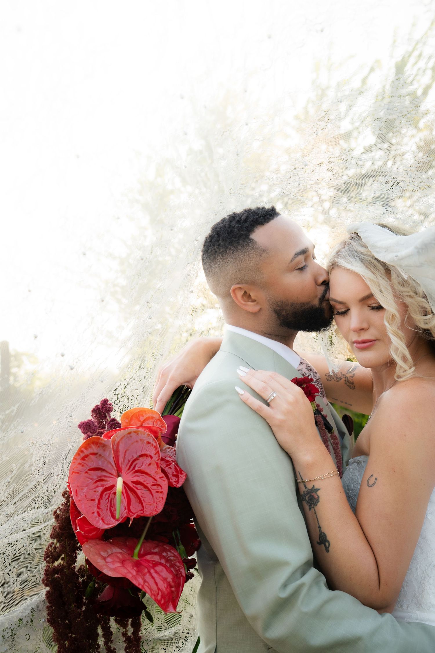 Groom kissing bride holding vibrant red anthurium bouquet with dramatic backlit floral backdrop.