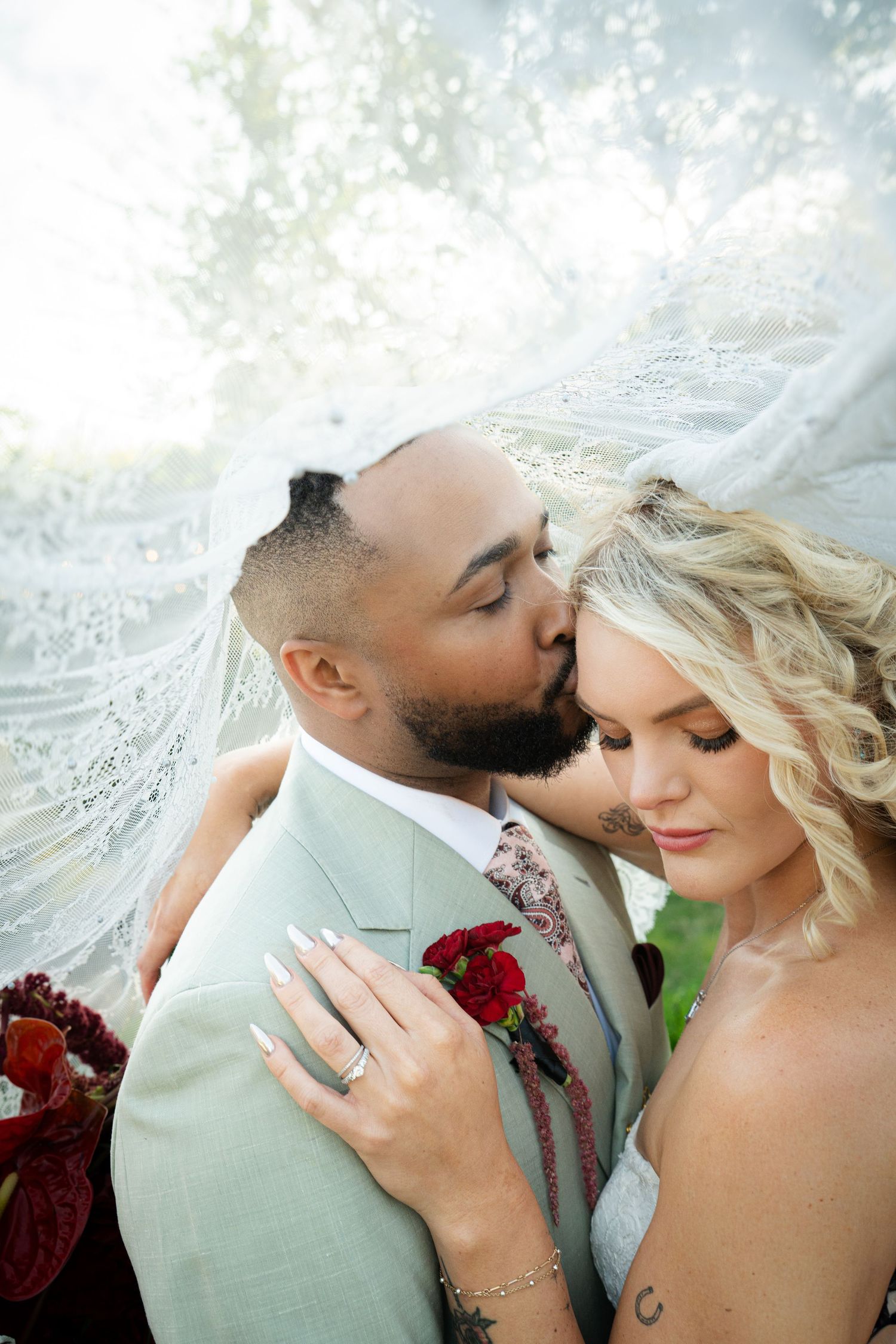 Groom in sage suit kisses bride's temple under flowing lace veil, bride holds red rose bouquet.