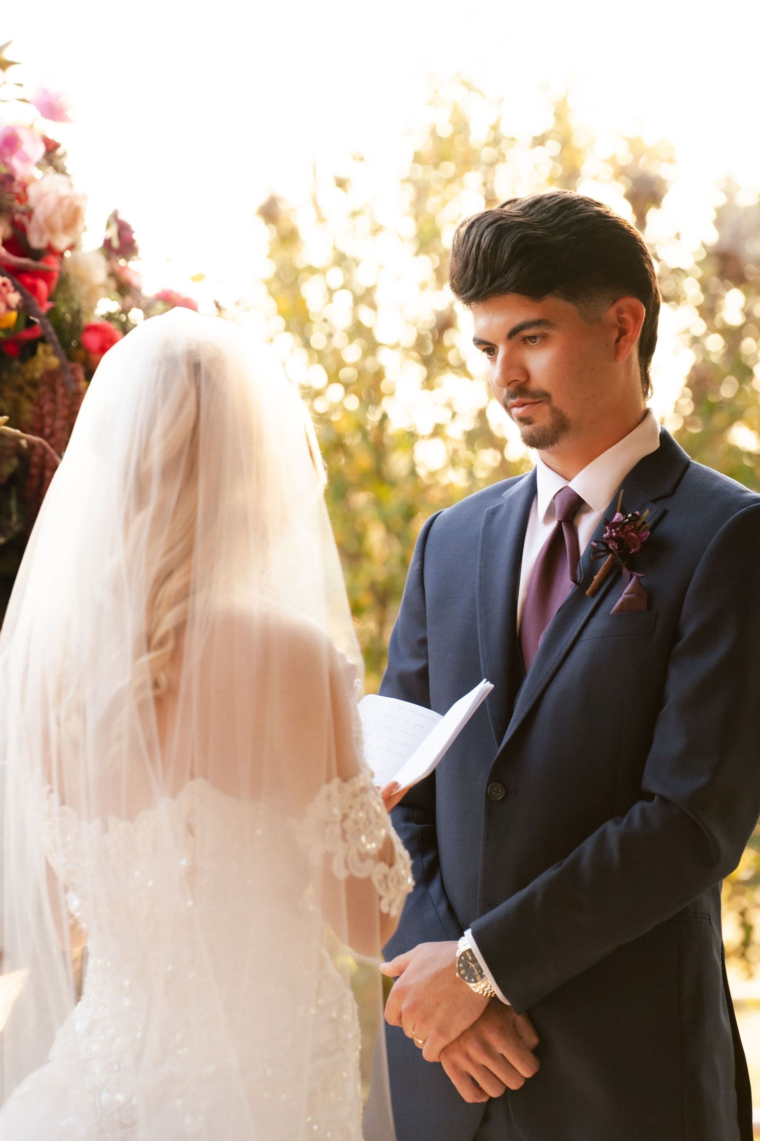Groom in navy suit reads vows to bride in lace gown at outdoor wedding ceremony.