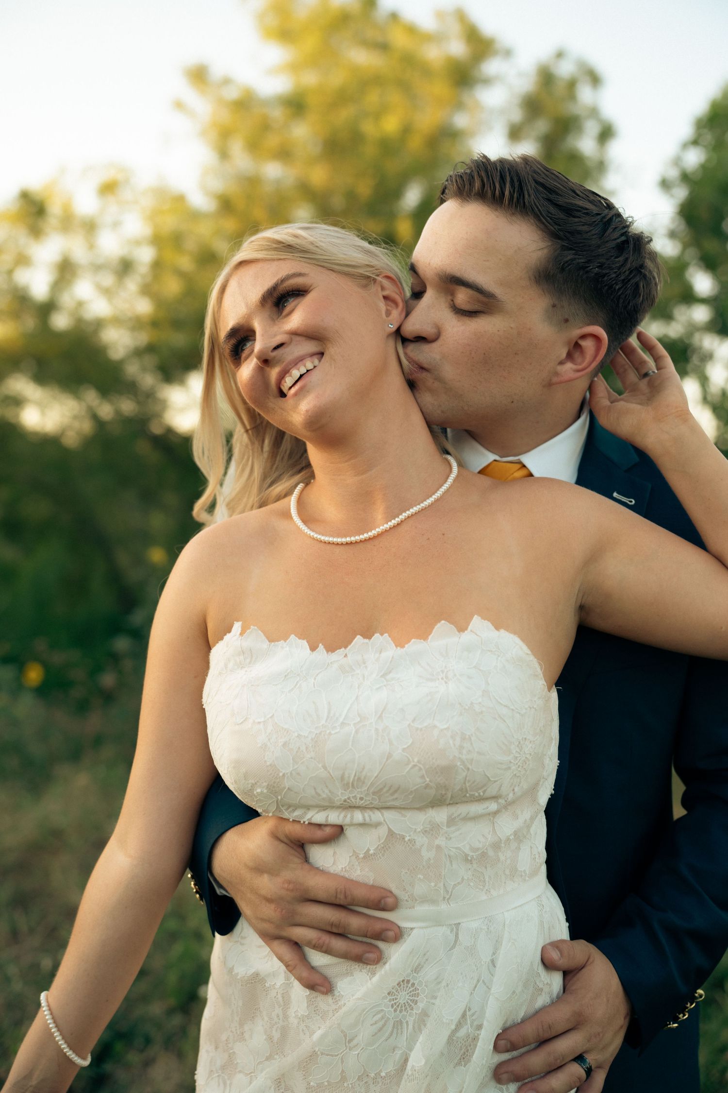 Groom in navy suit kisses bride on cheek as she smiles joyfully in white strapless lace wedding dress outdoors.