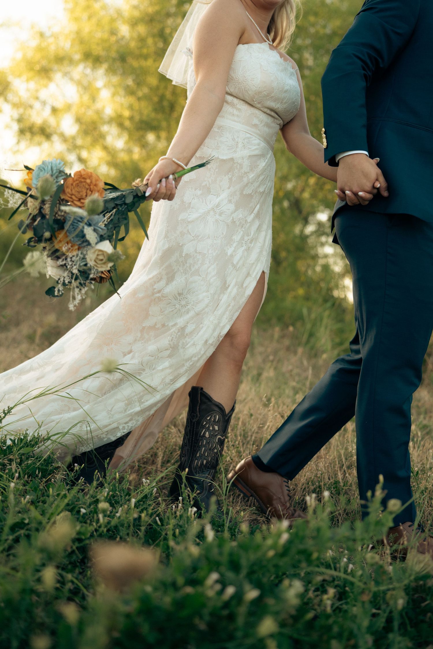 Bride in lace gown wearing cowboy boots walks holding bouquet hand in hand with groom outdoors.