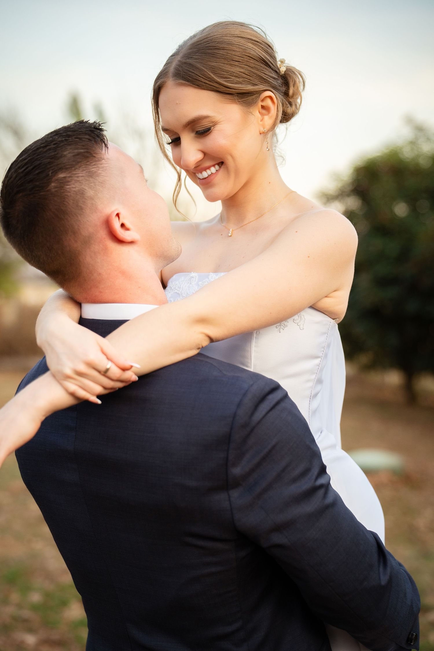Groom in navy suit lifts smiling bride in strapless white gown outdoors during golden hour wedding portraits.