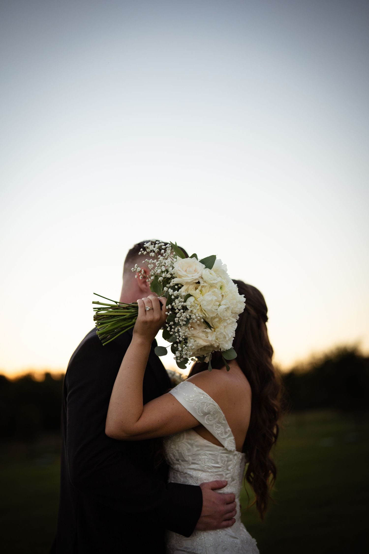 Bride and groom share a romantic kiss hidden behind a white rose bouquet at sunset.
