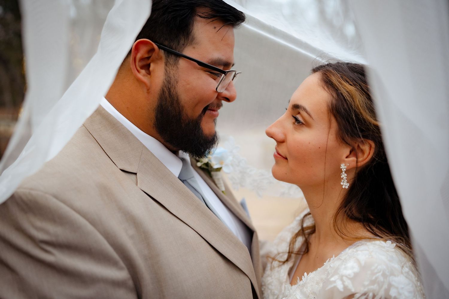 Bride and groom in tan suit share a romantic moment under a white veil outdoors.