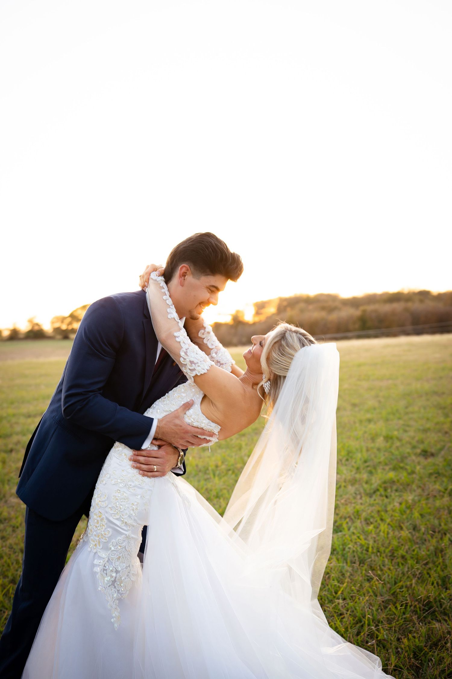 Bride and groom share a romantic dip kiss in a golden sunlit field, bride wearing lace gown and veil.