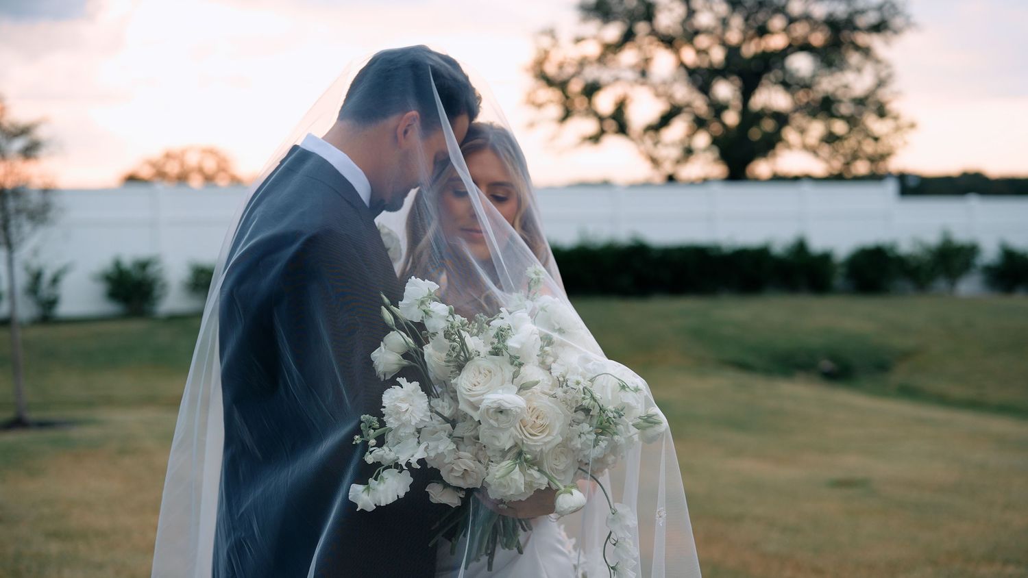 Bride and groom sharing romantic moment under flowing veil at sunset with white bouquet.