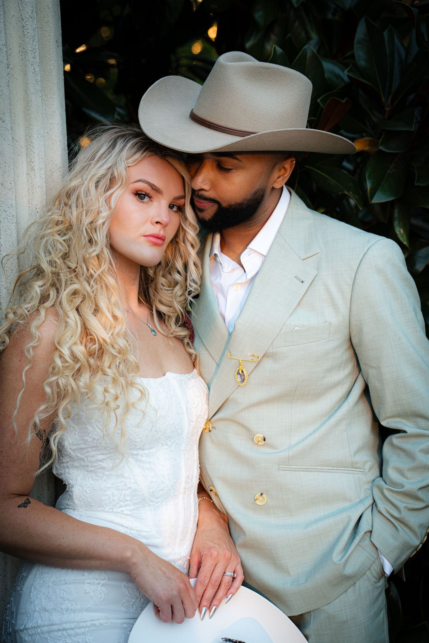 Elegant couple poses intimately with bride in white lace dress and groom wearing a tan cowboy hat and suit.