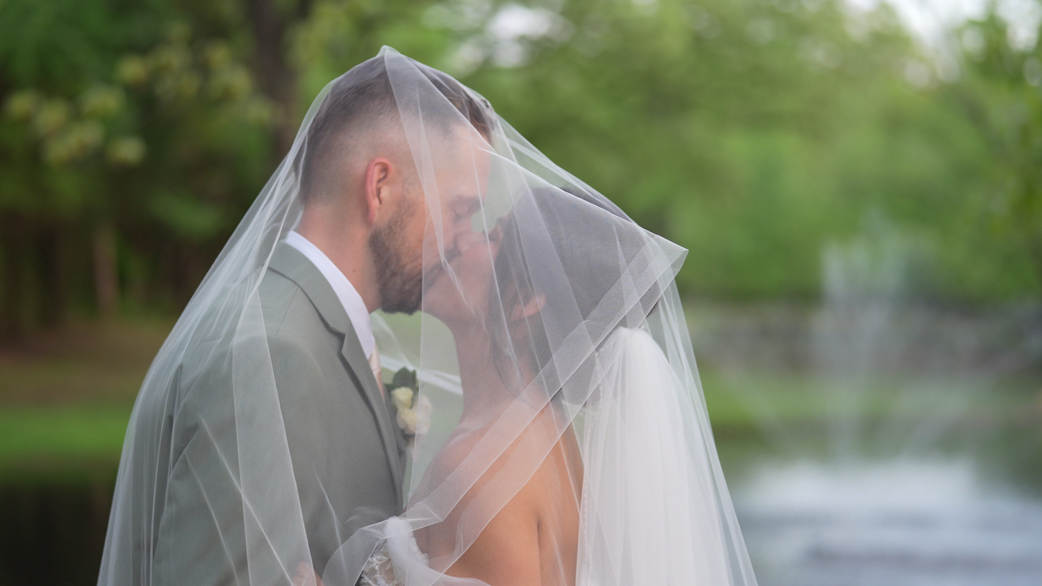 Bride and groom kissing under a veil near a serene lake.