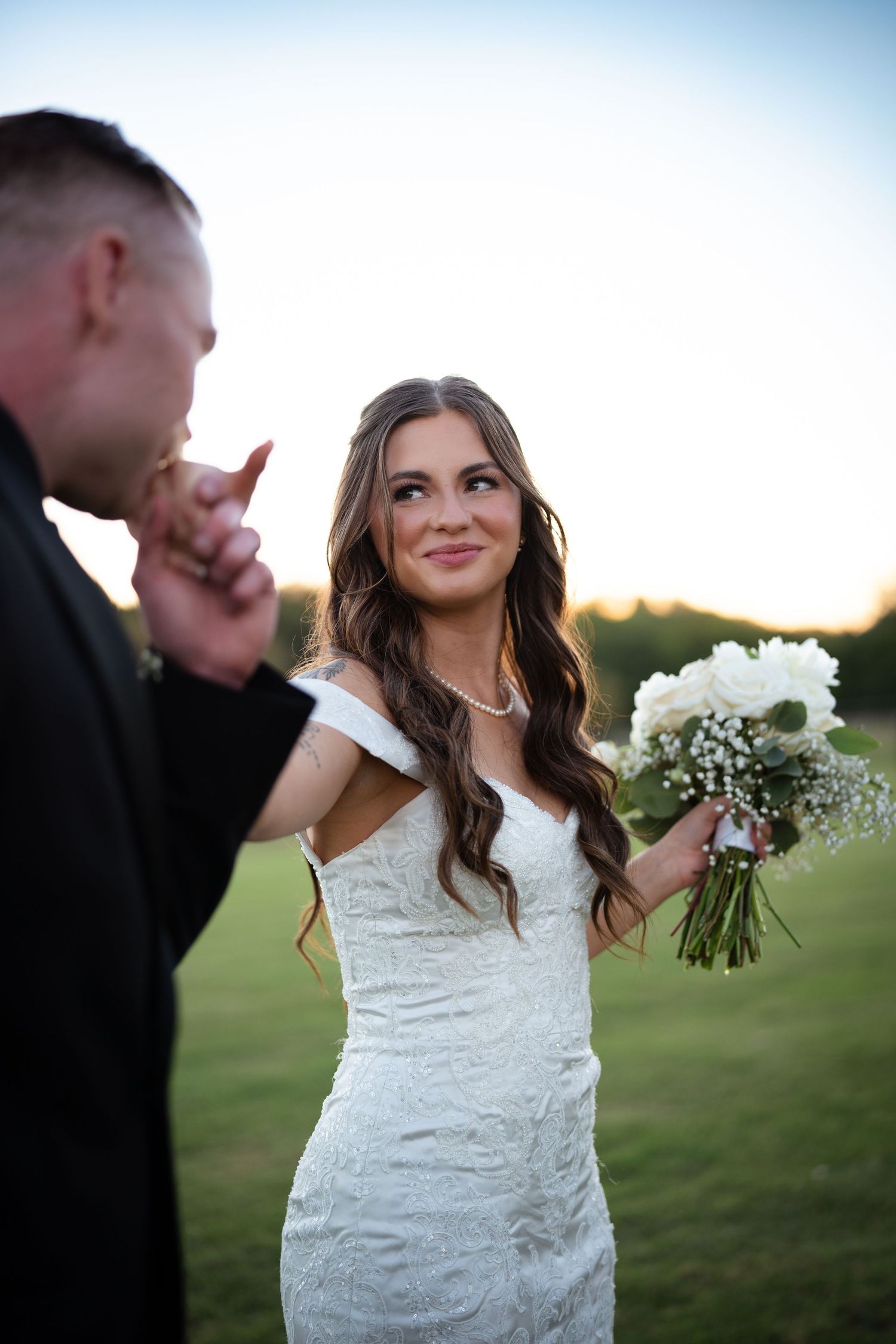 Radiant bride in off-shoulder lace gown smiles as groom kisses her hand holding white bouquet.
