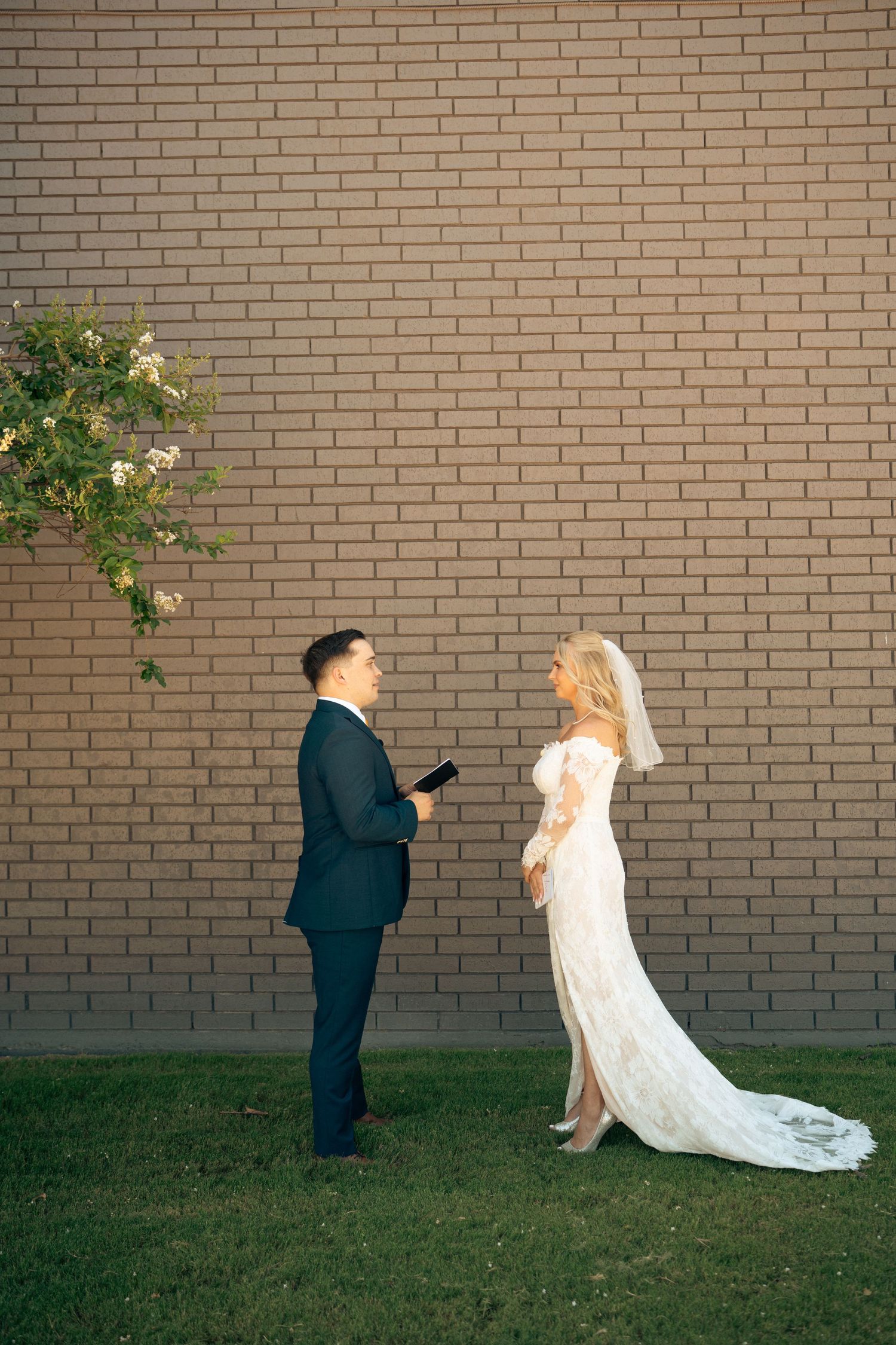 Groom reading vows to bride in white lace dress against brick wall outdoors.
