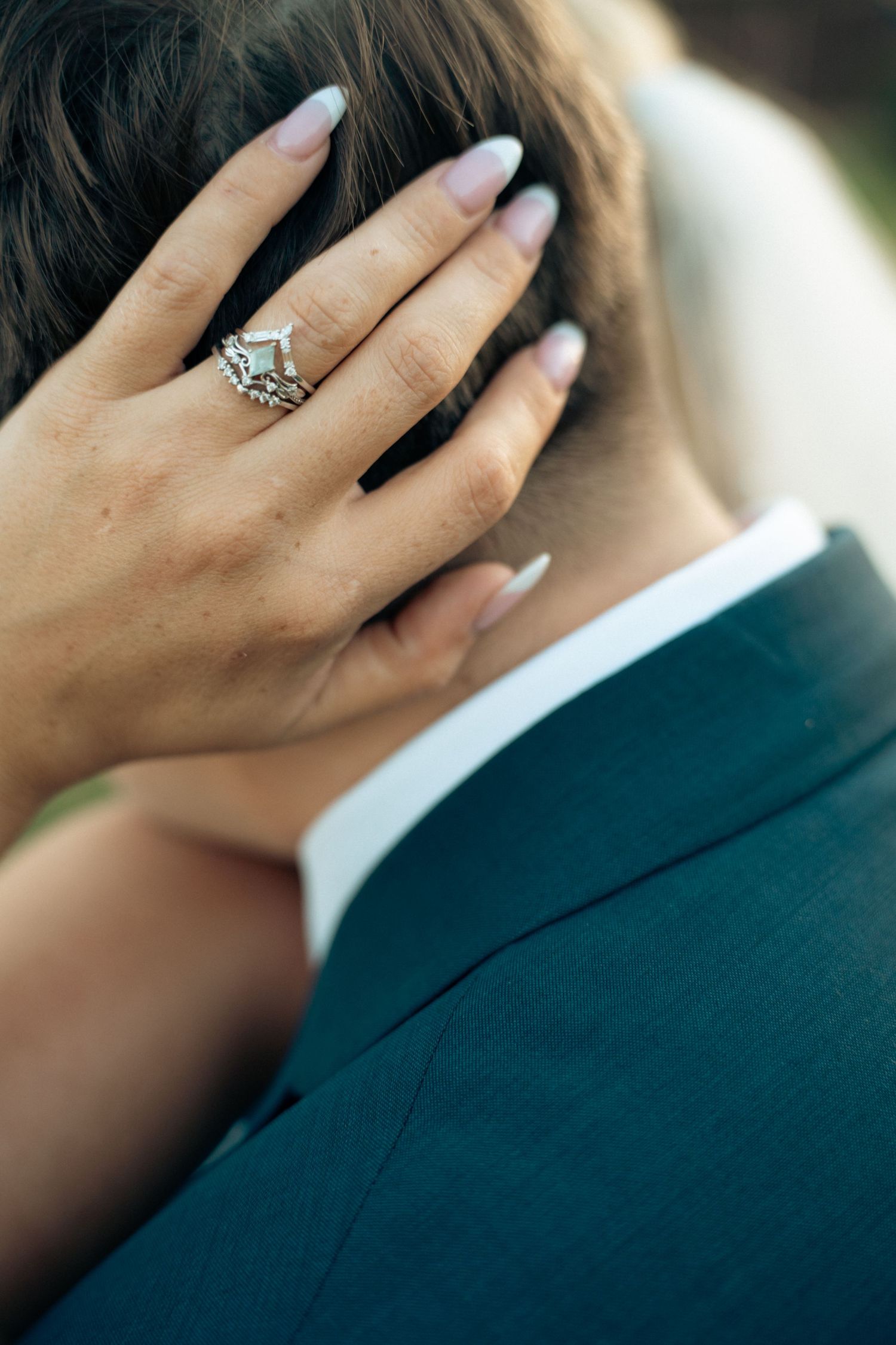 Close-up of bride's hand with diamond wedding ring set resting on groom's head in blue suit.