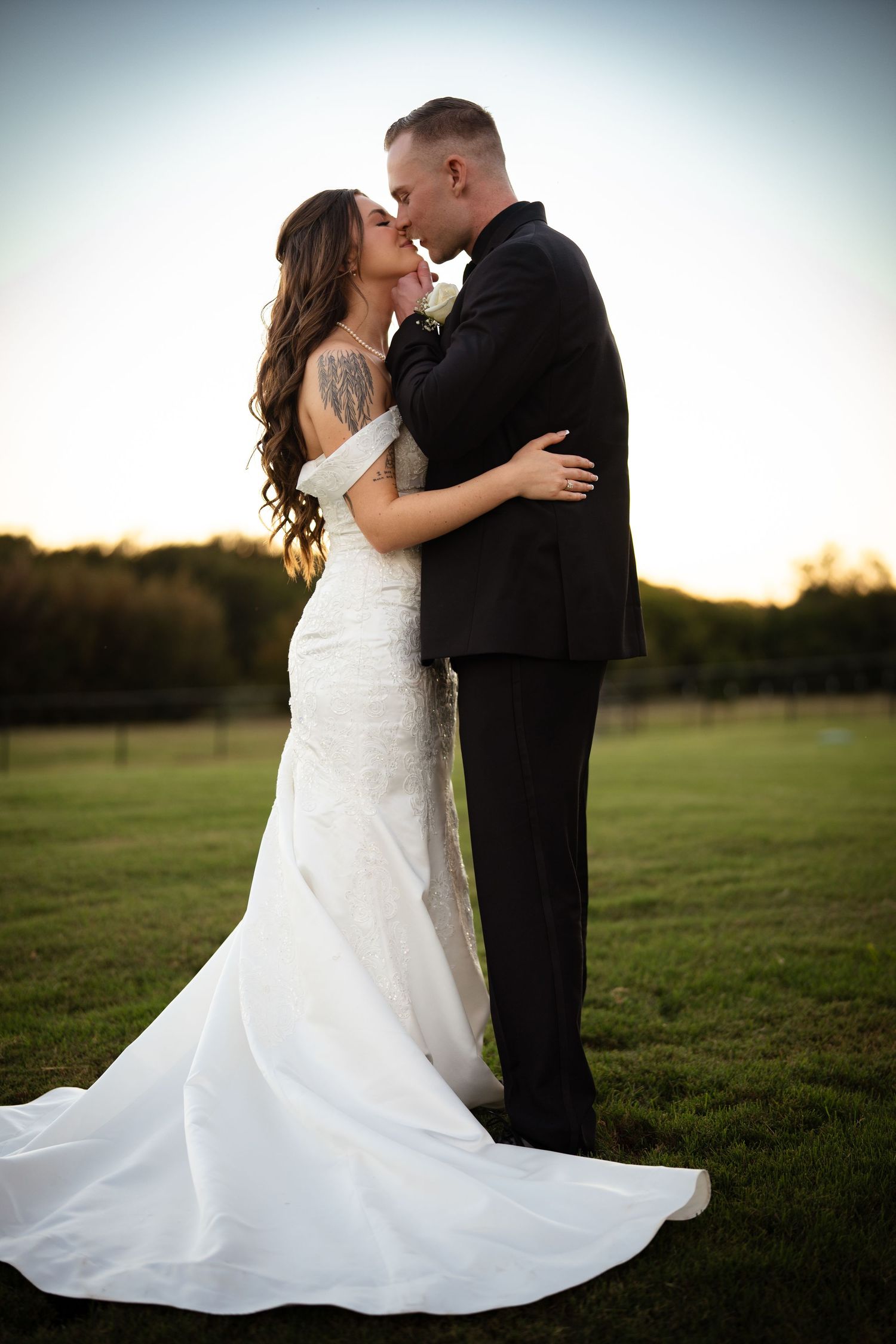 Bride in mermaid gown and groom in black suit share a tender kiss on a green field at sunset.