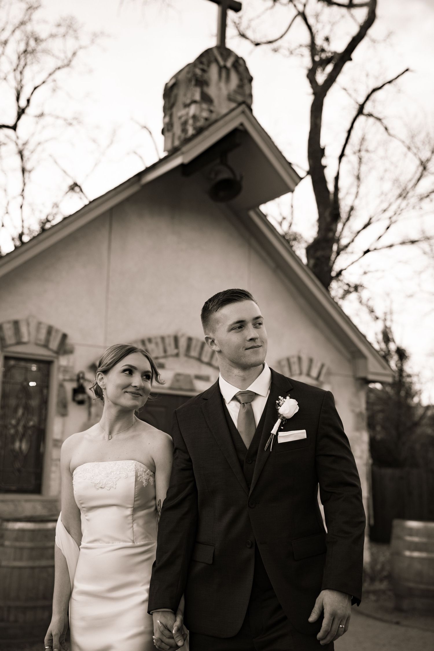 Elegant black and white wedding portrait of bride and groom holding hands before a rustic chapel.
