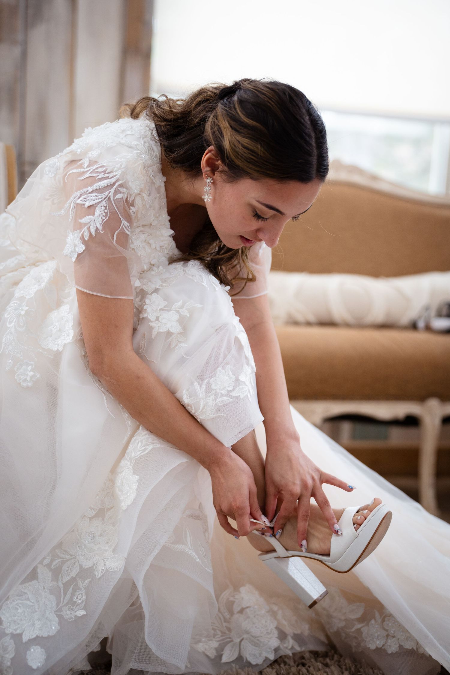 Bride in lace wedding gown sitting on floor fastening her white heeled bridal shoes.