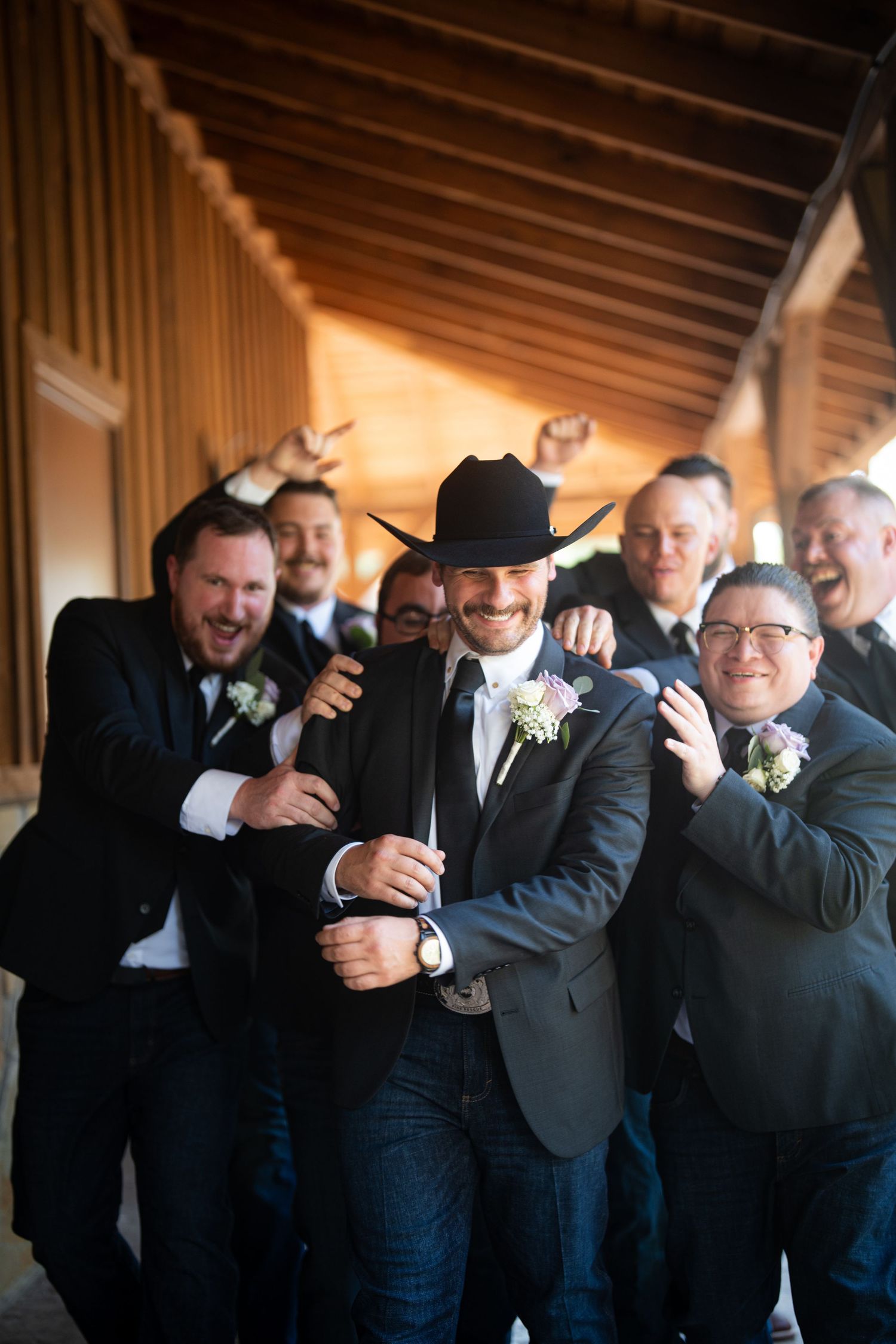 Groom in black cowboy hat and jeans celebrates with groomsmen in gray suits and boutonnieres at rustic wedding venue.