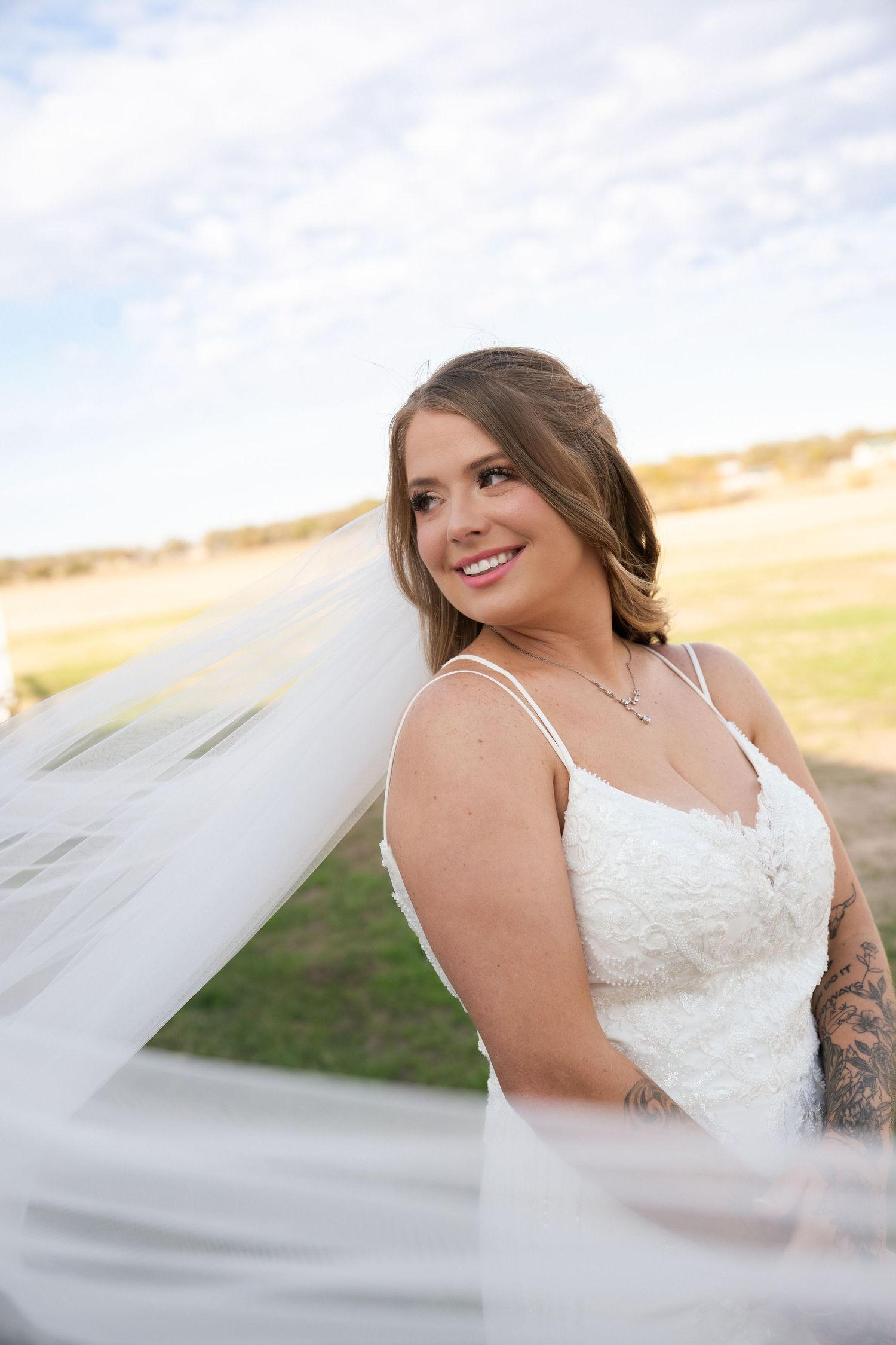 Smiling bride in lace gown with flowing veil poses outdoors under a partly cloudy sky.