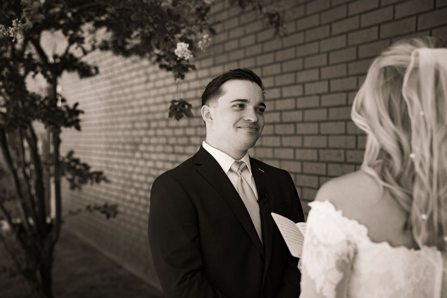 Black and white photo of groom smiling warmly at bride during intimate vow exchange outdoors.