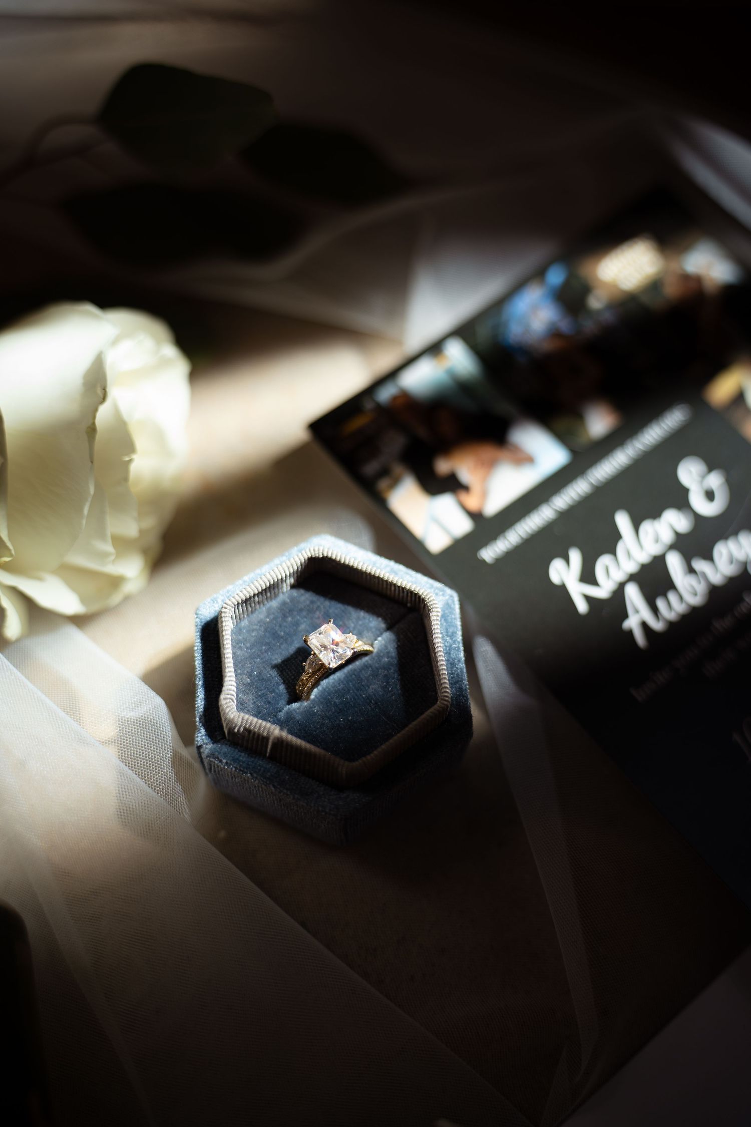 Diamond engagement ring in gray velvet box beside wedding invitation for Kaden and Aubrey and white rose.