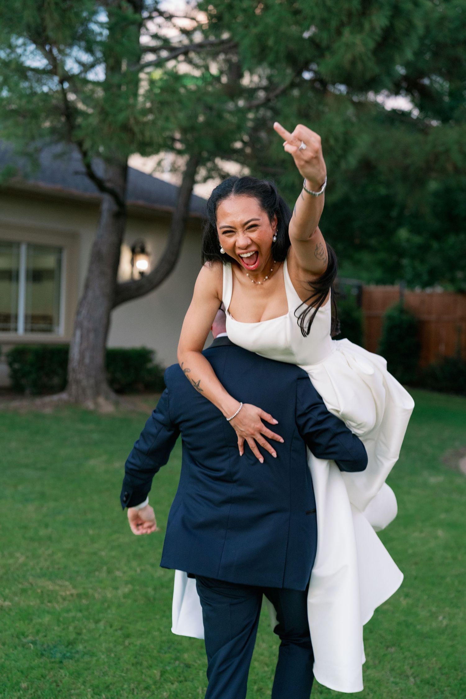 Groom carrying excited bride on his back across green lawn at outdoor wedding reception.